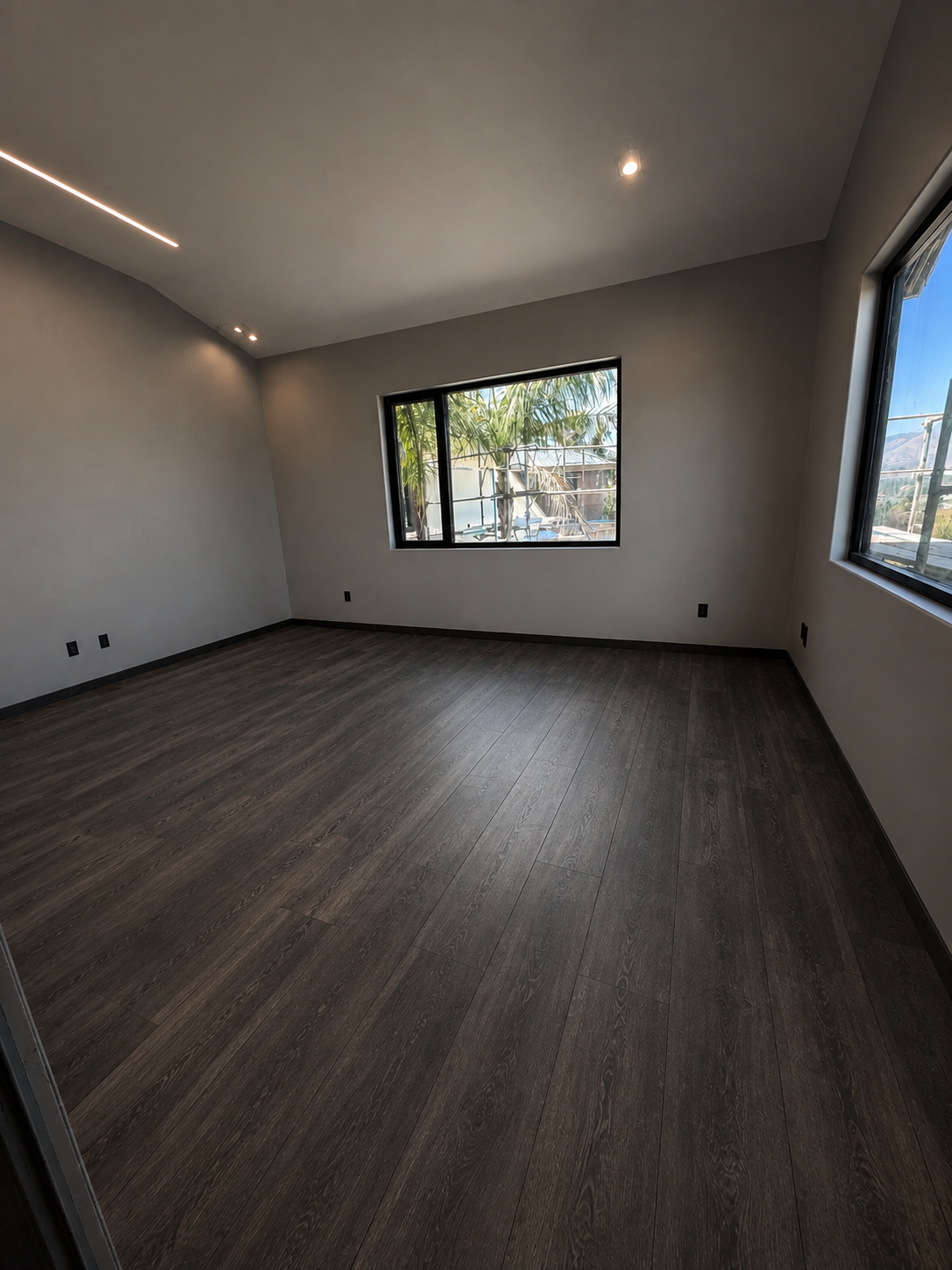 Empty room with dark wood floors, white walls, and large windows showing palm trees outside.