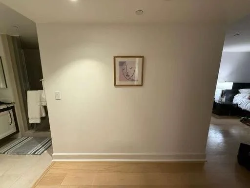 A blank wall with a framed line drawing of a woman's face hanging on it, in a hotel room or apartment, with a partial view of a bed and a bathroom in the background.