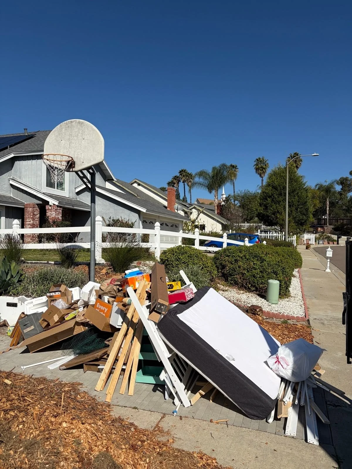Overflow of discarded items and debris on the sidewalk in front of a residential neighborhood, including cardboard boxes, wood pieces, and a large unfolded mattress, with houses, palm trees, and blue sky in the background.