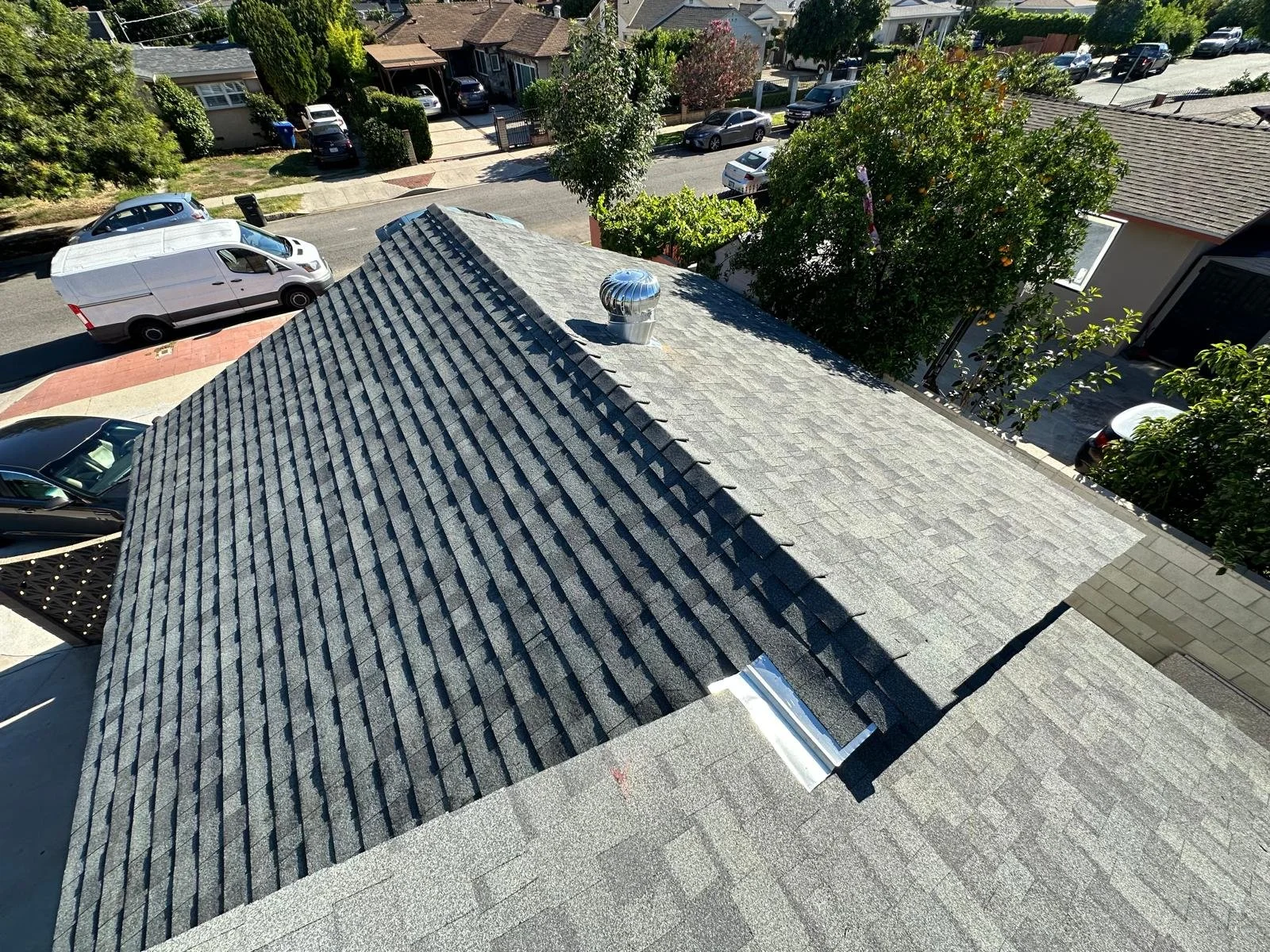 Aerial view of a residential neighborhood with parked cars, trees, and houses, focusing on a house's roof with gray shingles and a metal vent.