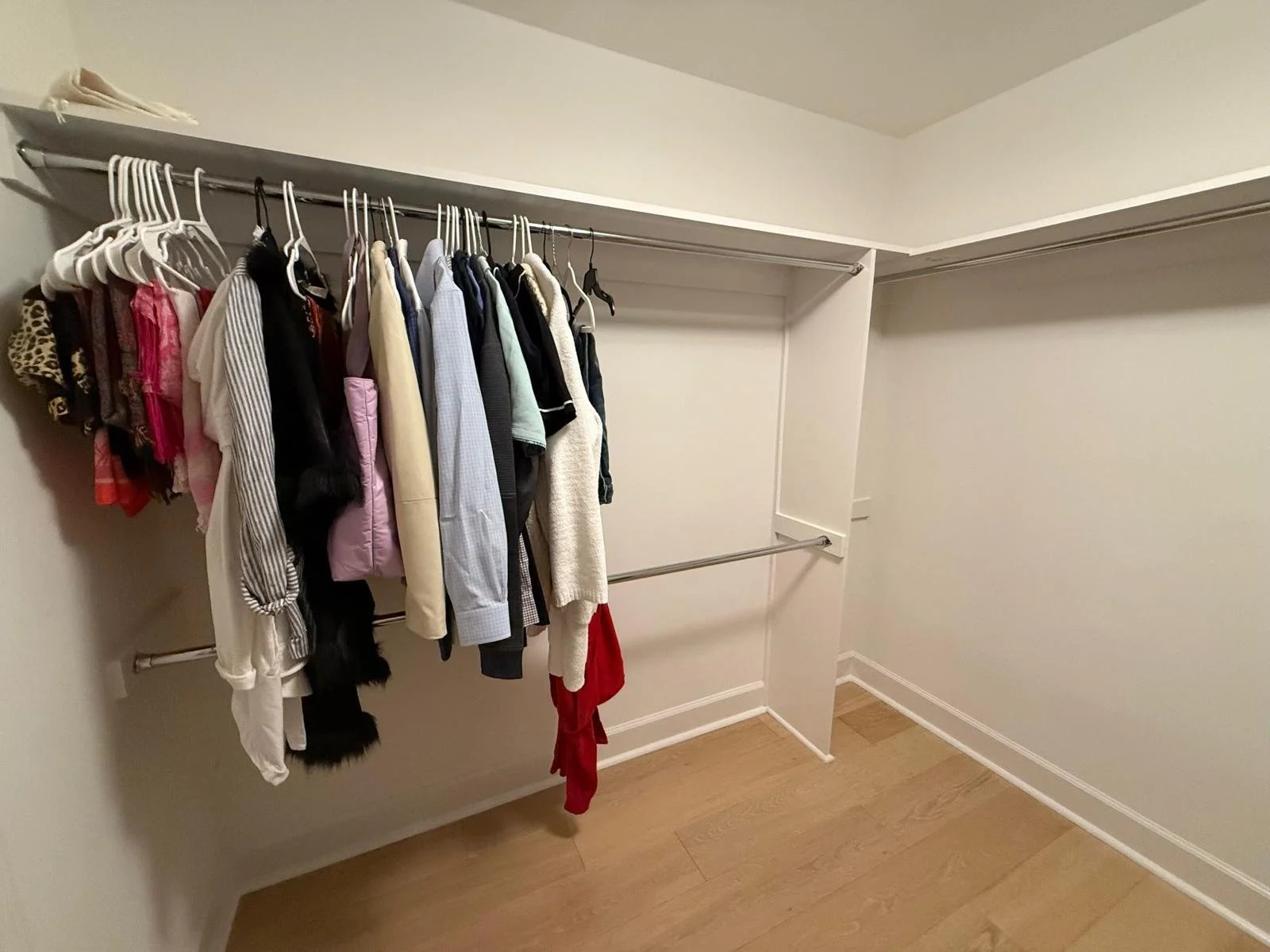 Empty walk-in closet with clothes hanging on metal rods, white walls, and wood flooring.