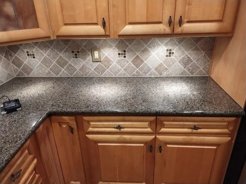 Kitchen countertop with wooden cabinets above and below, tiled backsplash, and an electrical outlet.