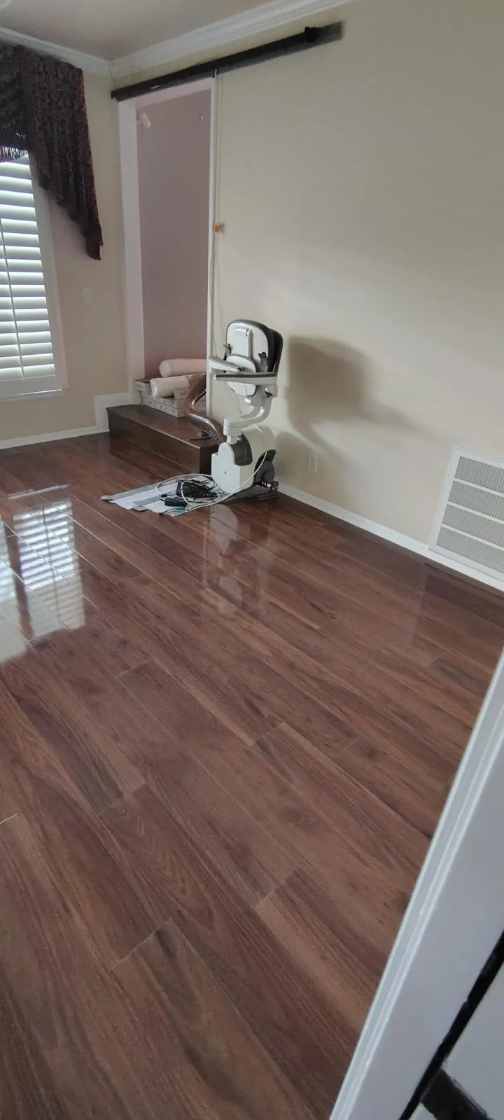Empty room with polished wood floor, a wall-mounted medical or technical device, cords, and a window with blinds and curtains.