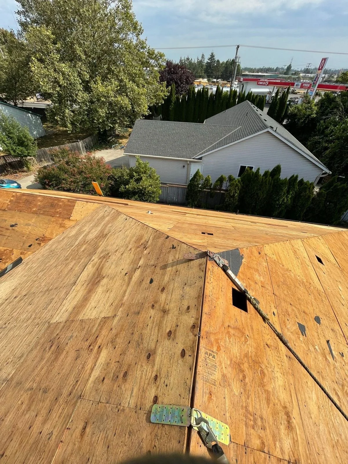 View from a rooftop under construction showing plywood sheathing, a nearby residential house with a gray roof, green trees, a parking area, and a commercial area in the distance.