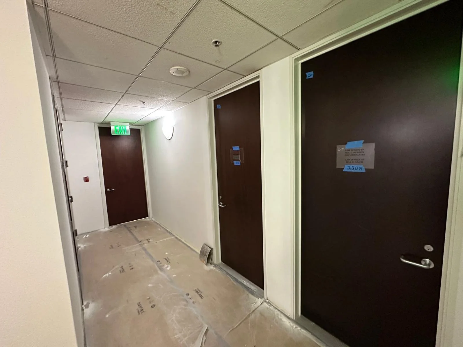 Hallway with beige walls, brown doors, and ceiling tiles. One door is labeled with a blue tag. The floor is covered with protective plastic sheeting. An illuminated green exit sign is visible above one door, and a wall-mounted light is turned on.