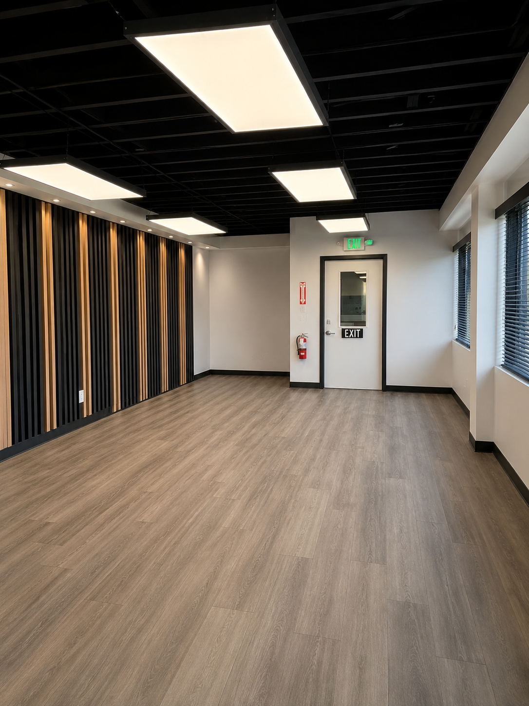 Empty modern room with wood flooring, black ceiling with rectangular white lights, and a wall with vertical black and wood panels. There is a door labeled 'EXIT' with an emergency exit sign above, a fire extinguisher on the wall, and windows with bli
