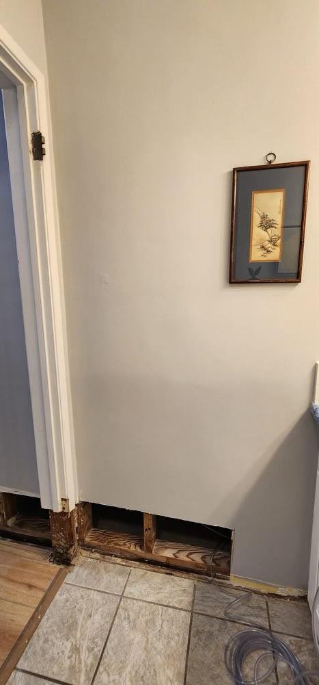 Interior wall with a framed picture, missing baseboard, and exposed wooden foundation beneath a cut-out section. Part of a door frame is visible on the left.