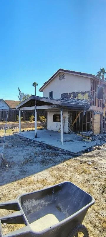 Under construction house with a small covered patio, unfinished walls, and a wheelbarrow in the foreground under a clear blue sky.