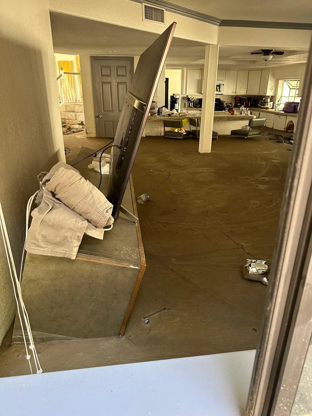 Living room and kitchen area in a house with damaged flooring and scattered belongings, including a computer monitor on a table, clothes, water bottles, and small items on the counters.
