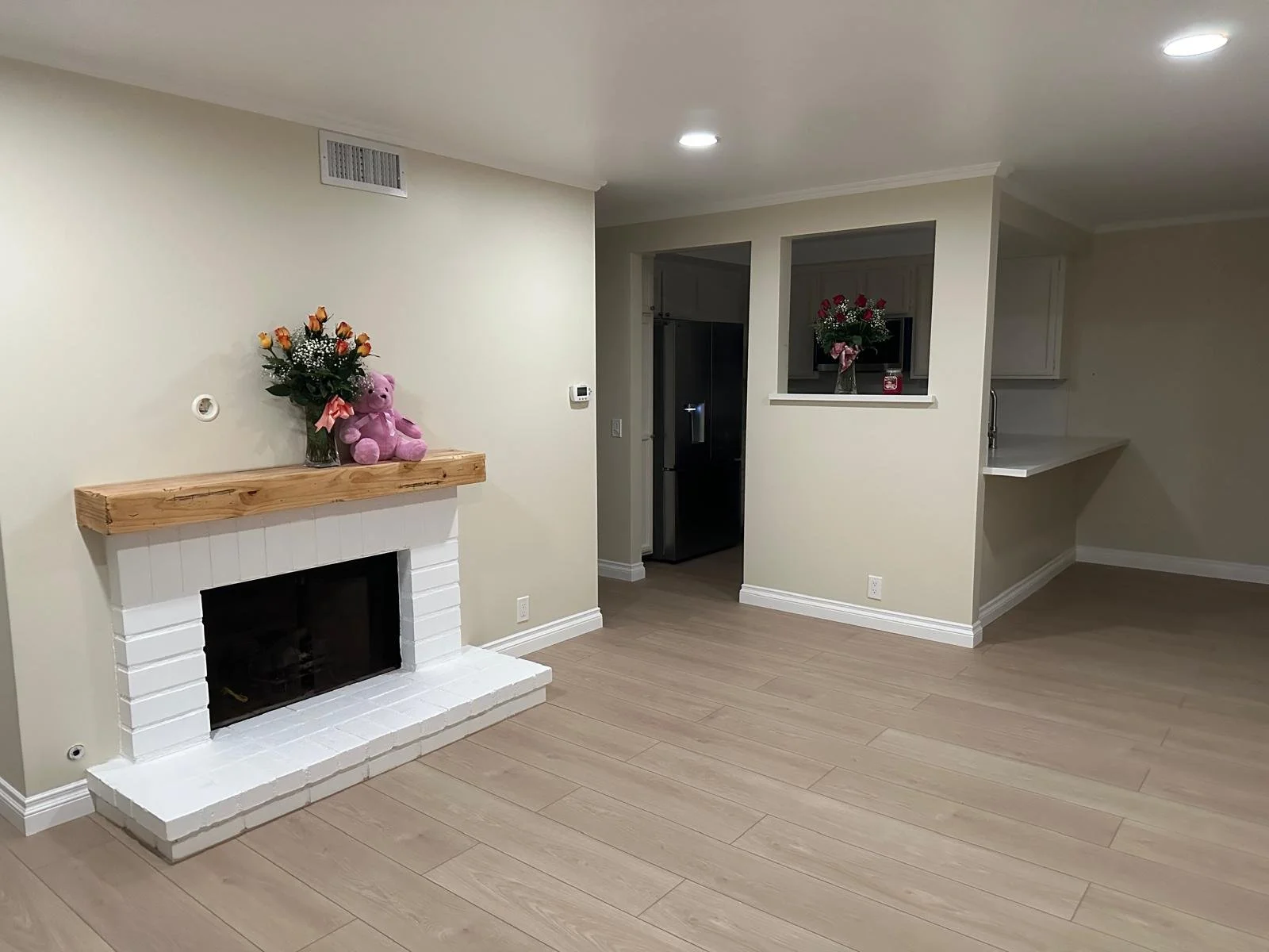 Empty living room with a white brick fireplace topped with a wooden mantel, decorated with a bouquet of roses and a pink teddy bear. The room has beige walls, light hardwood flooring, and a ceiling-mounted light. There is an open doorway leading to t