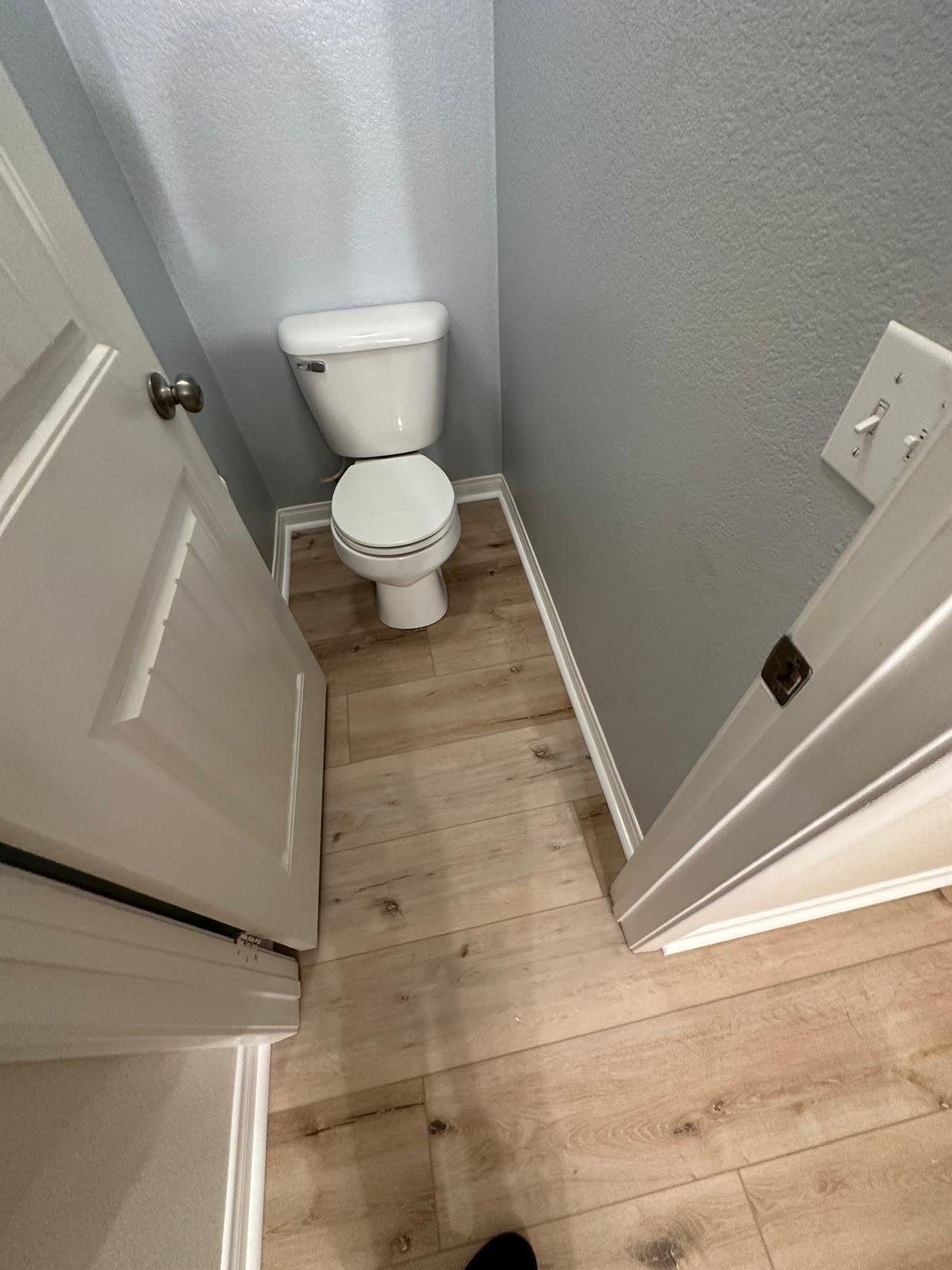 Small bathroom with a white toilet, gray walls, wood-like floor, and a door partially open.