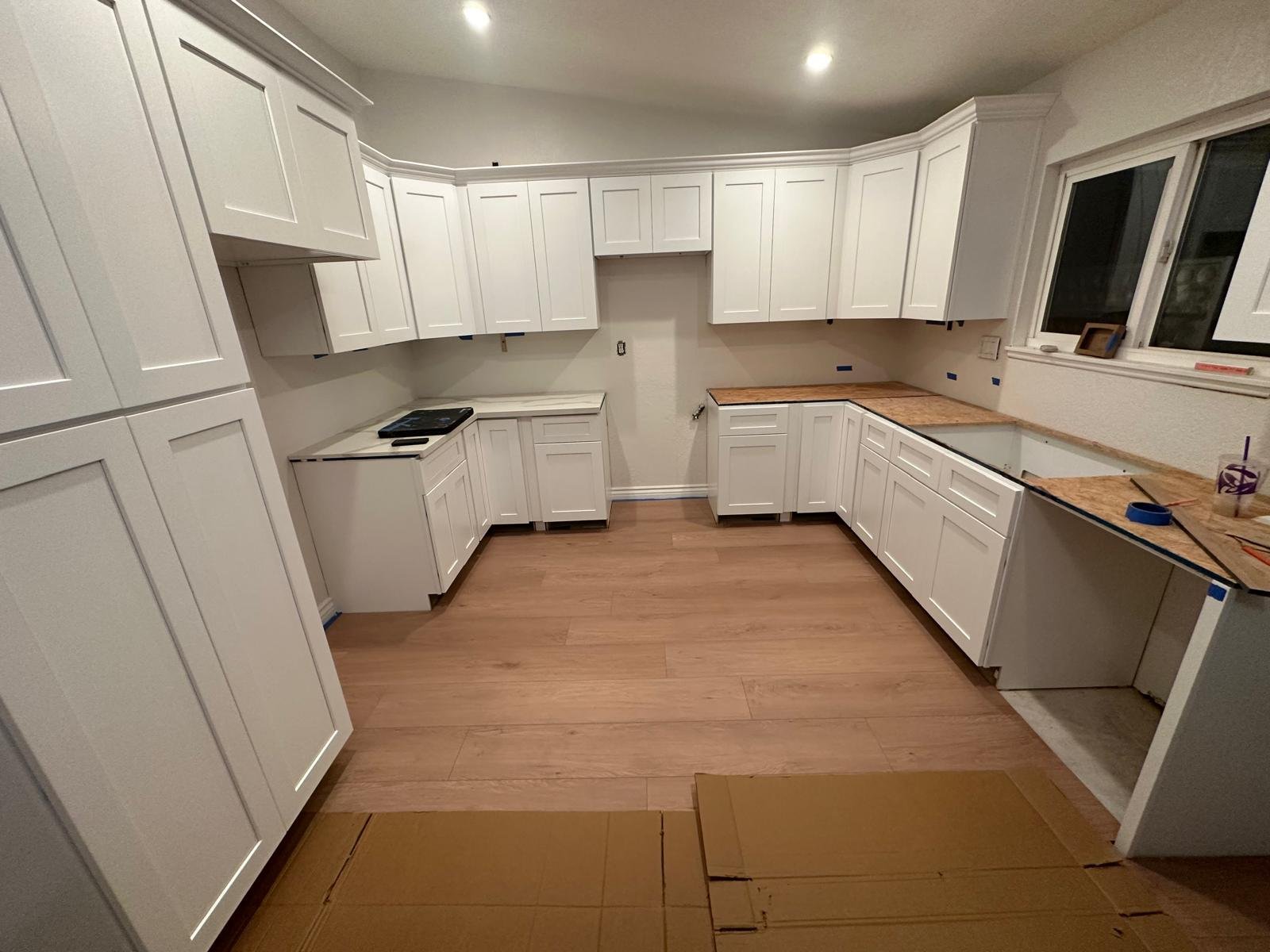 Kitchen with white cabinets, some with open space, and wood countertops. Some countertops are still under construction with tools and tape visible. The floor is wood, and a window is present on the right side.