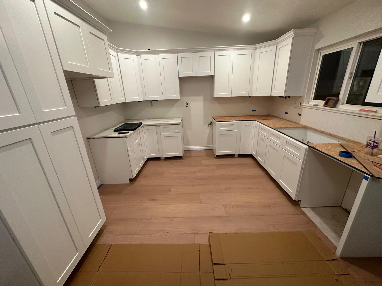 Kitchen with white cabinets, some with open space, and wood countertops. Some countertops are still under construction with tools and tape visible. The floor is wood, and a window is present on the right side.