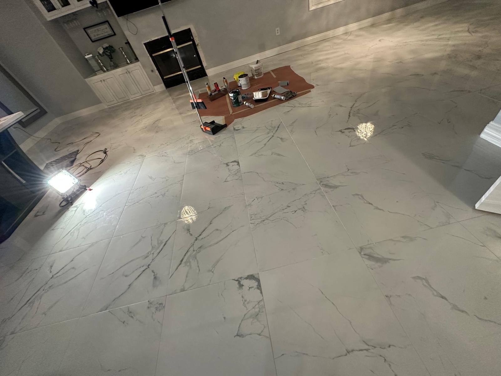 A room with marble tile flooring, tools, paints, and supplies arranged on a paper in the center, and construction lighting turned on. Walls are painted gray, with a built-in cabinet and fireplace in the background.