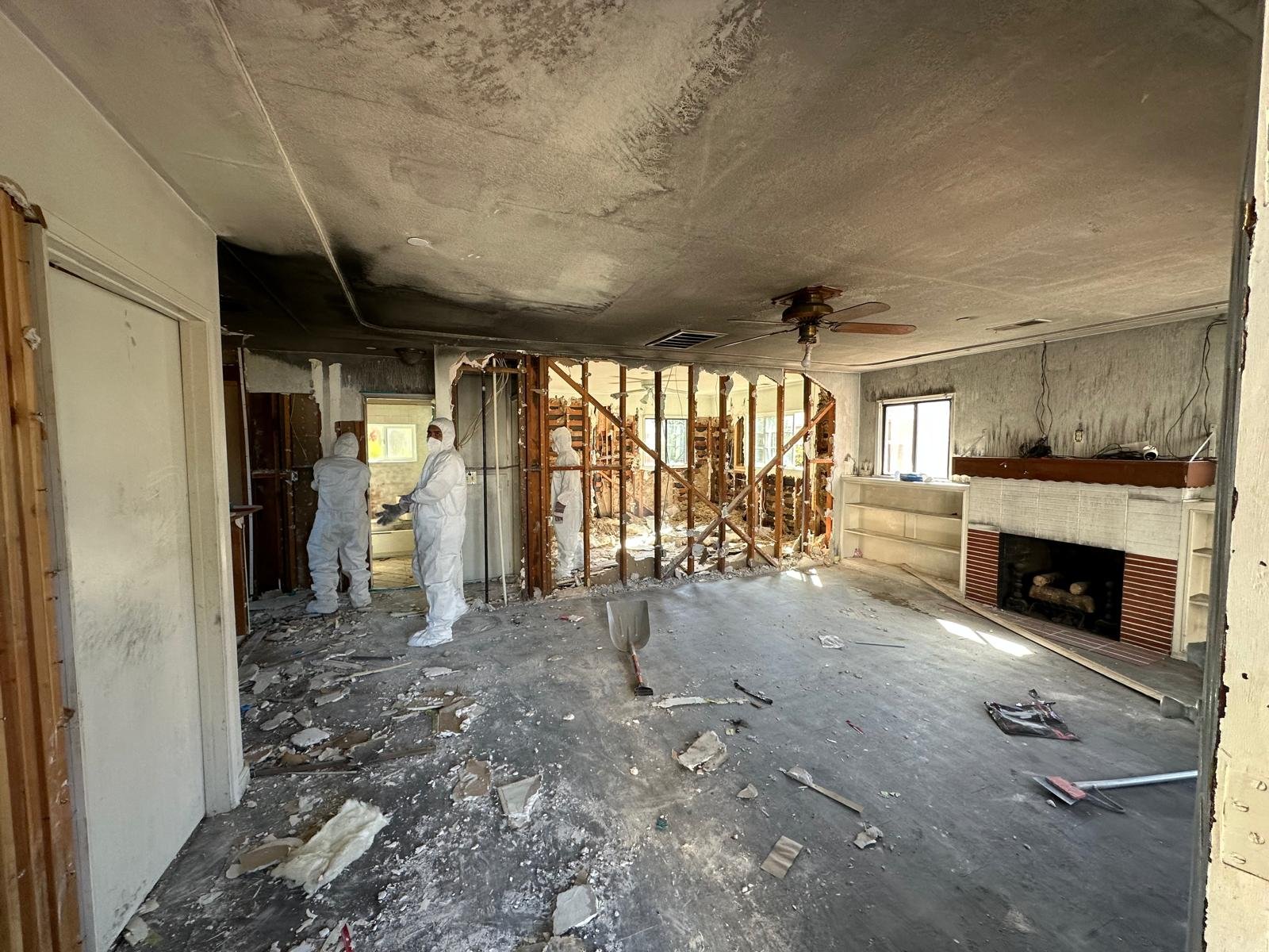The image shows a living room undergoing demolition with three workers dressed in white protective suits. The walls are partially torn down, exposing wooden studs. Debris is scattered across the floor, and a shovel is standing upright in the foregrou