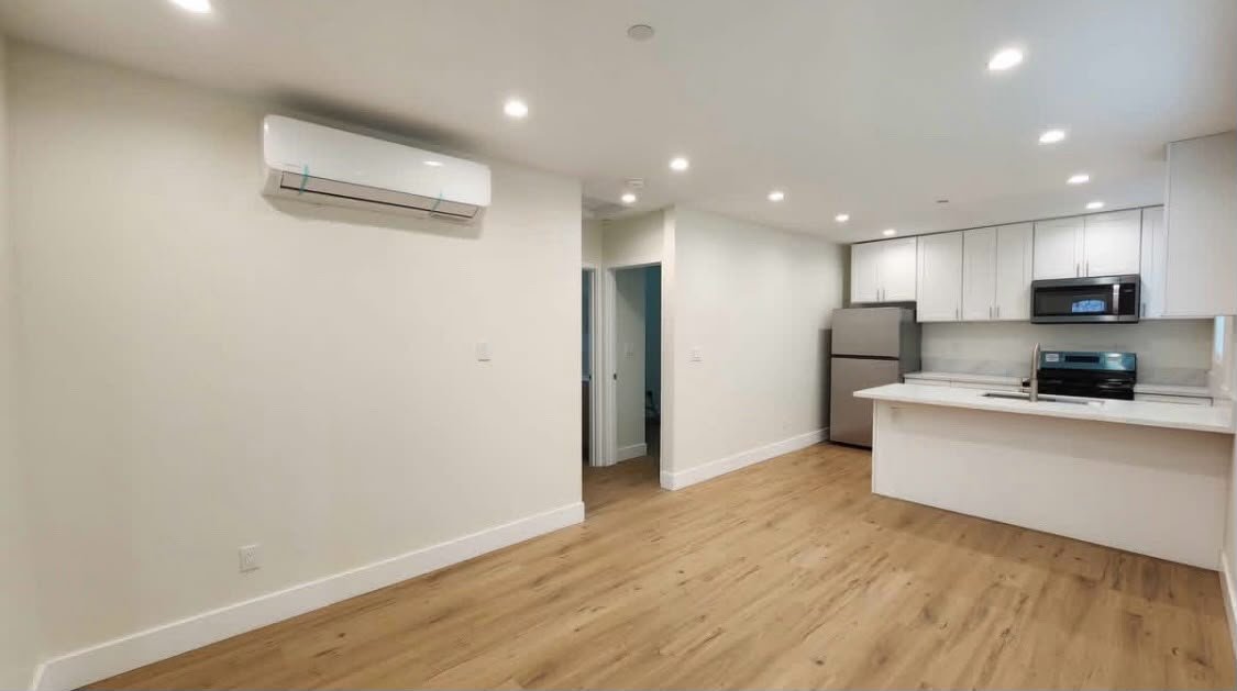 Empty living room and kitchen with white walls, hardwood floors, a refrigerator, microwave, and stove in a modern apartment.