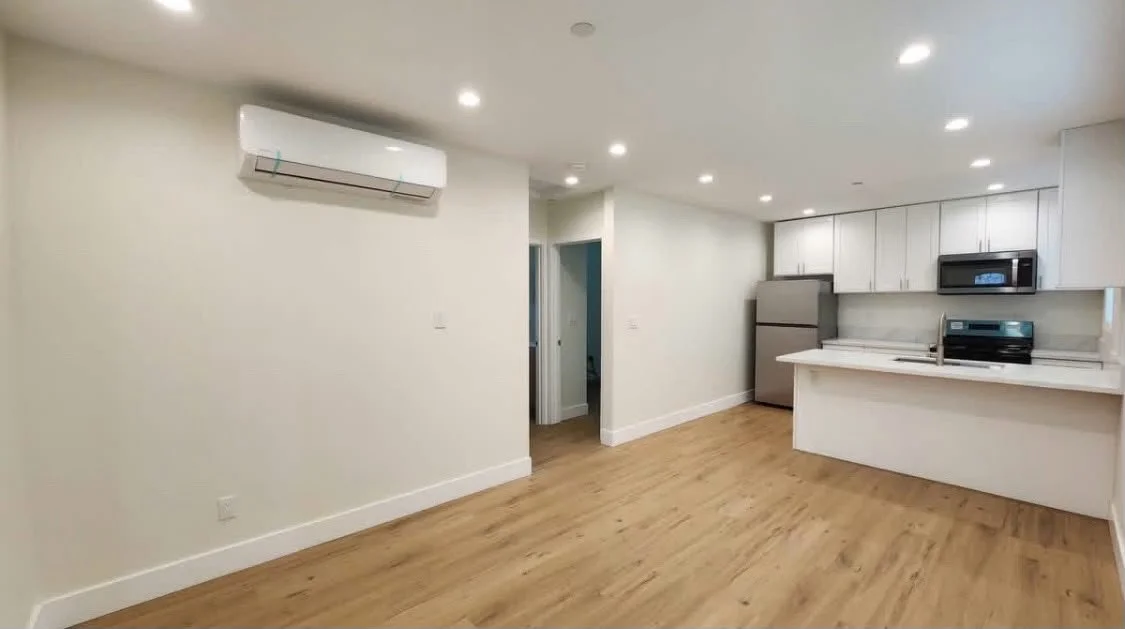 Empty living room and kitchen with white walls, hardwood floors, a refrigerator, microwave, and stove in a modern apartment.