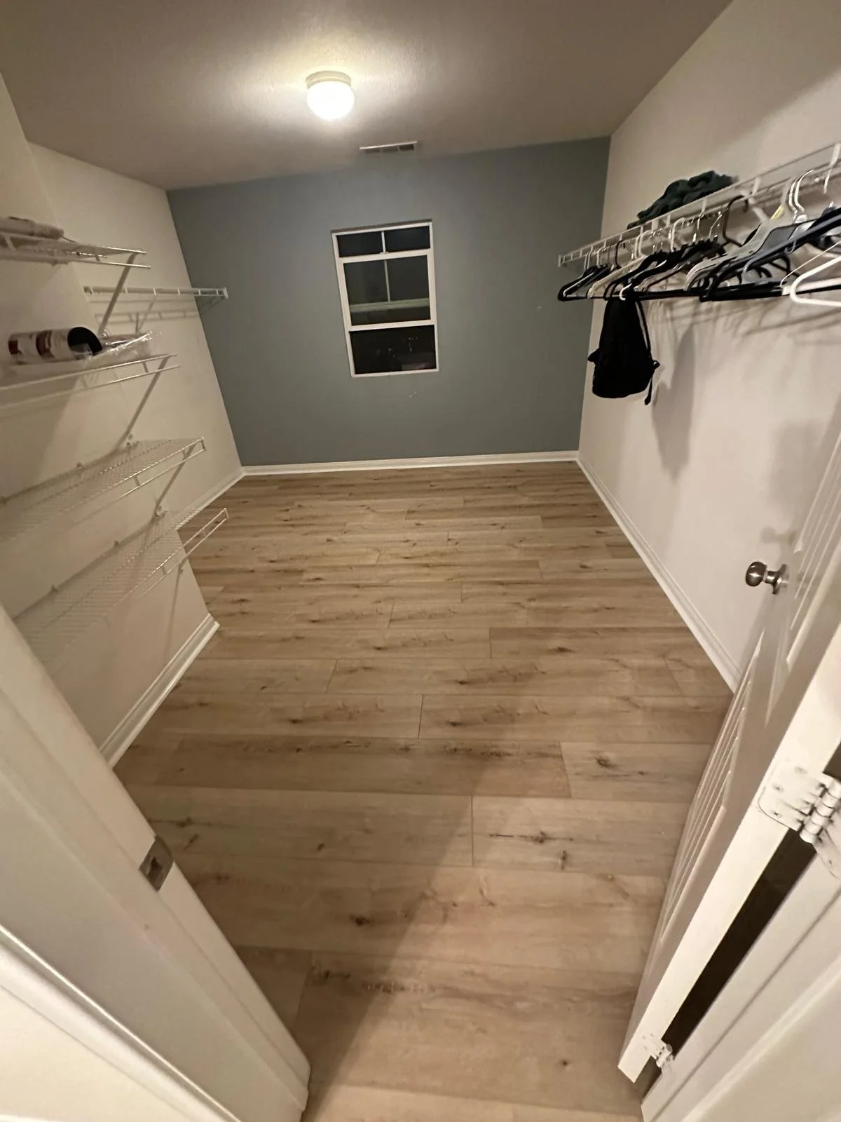 Empty walk-in closet with wood flooring, white walls, wire shelves on the left, and a window on the back wall.
