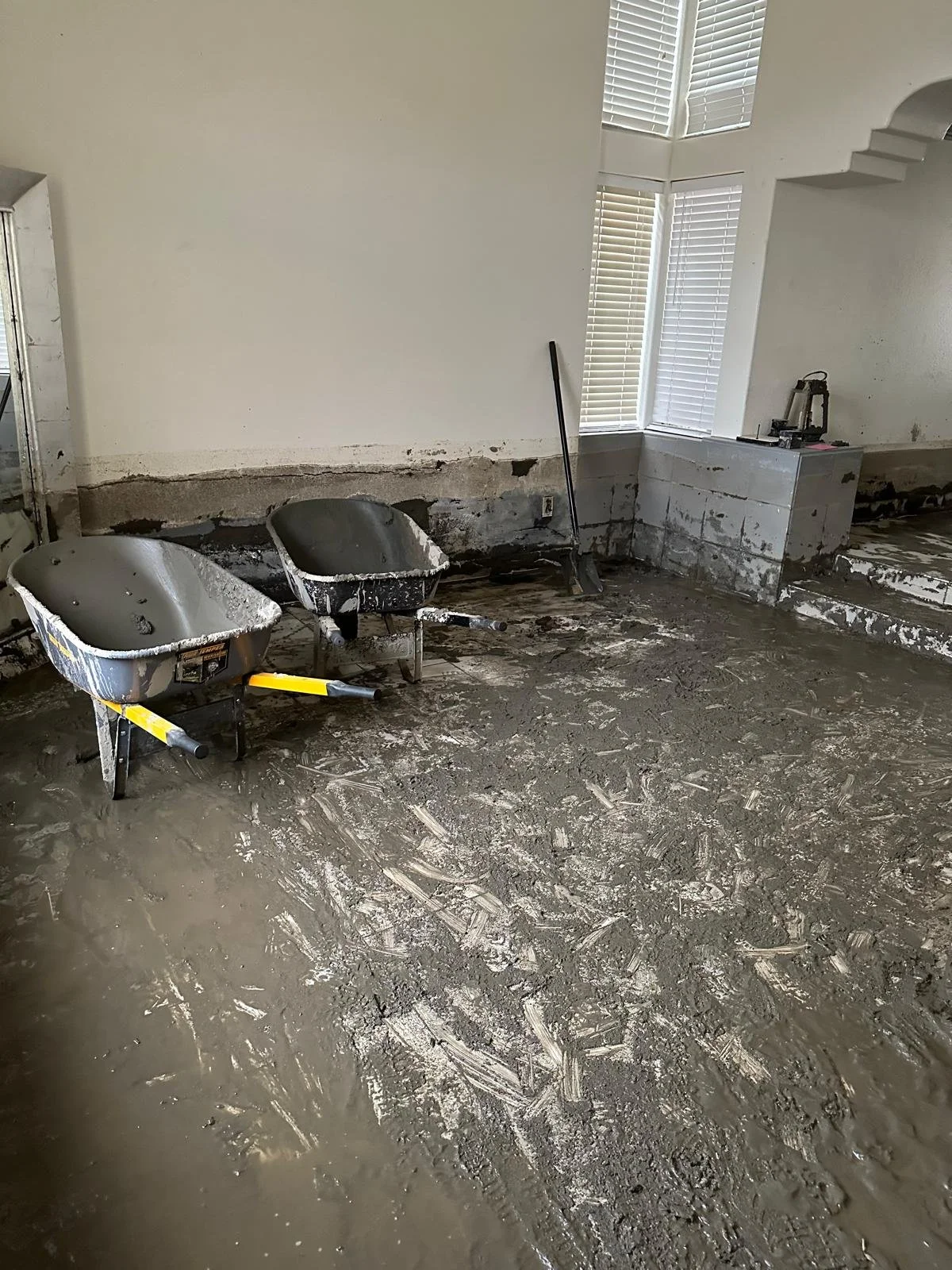 A room under renovation with wet cement on the floor, two wheelbarrows, a broom, and construction tools near a wall with partially removed drywall.