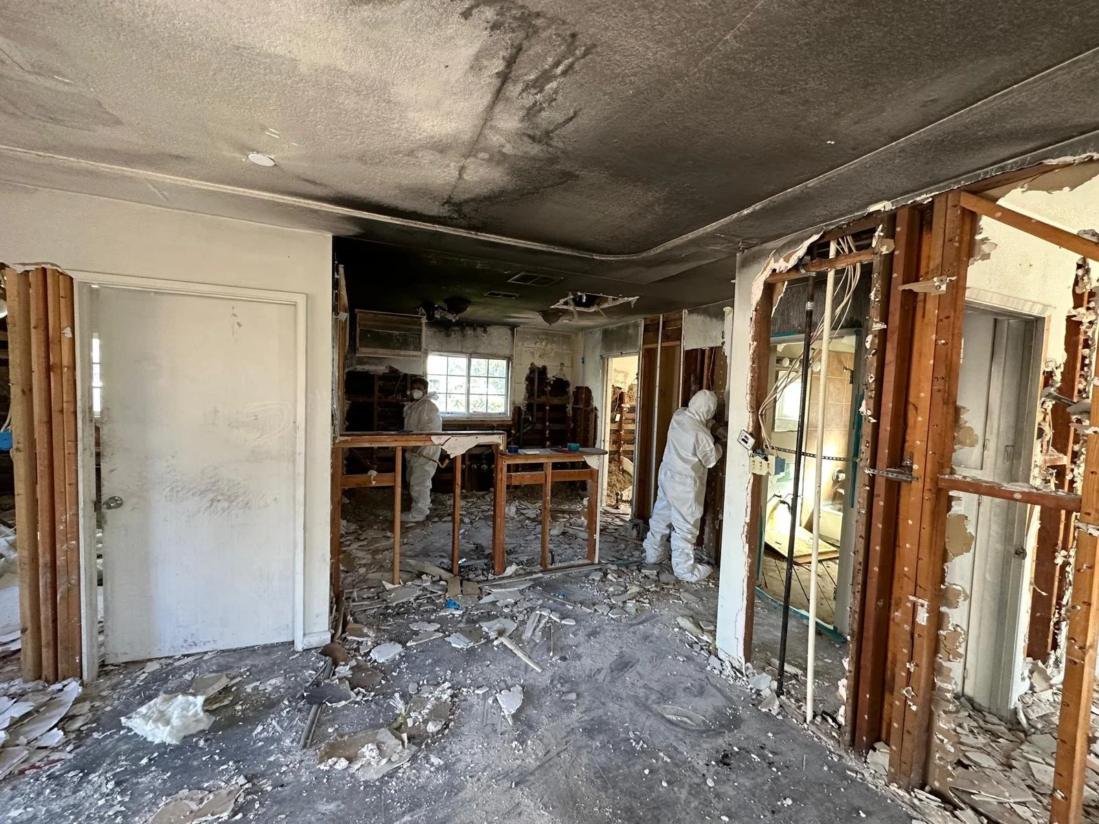 The image shows a room under renovation with drywall partially removed, exposing wooden framing and electrical wiring. Two workers in white protective suits are working in the room, which has a floor covered in debris and dust. The background feature