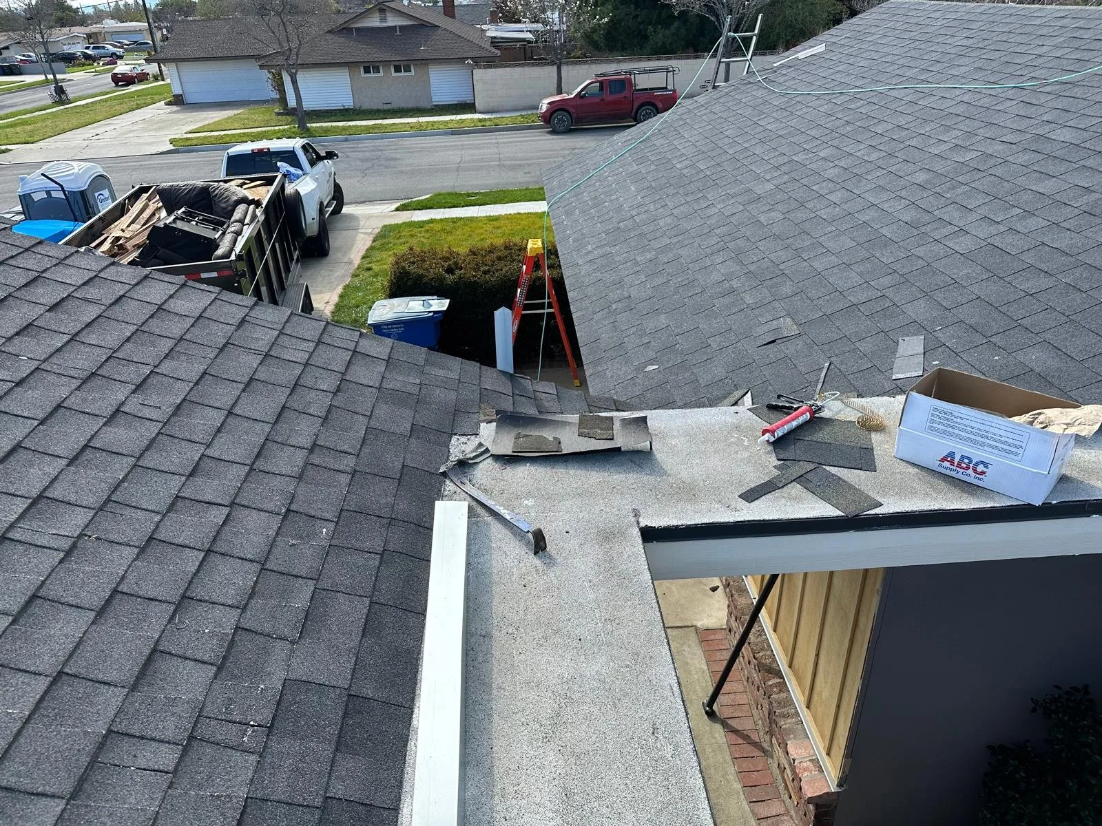 Roof with shingles undergoing repair, tools and materials scattered on the surface, ladder, vehicles parked nearby, and a street in front of the house.