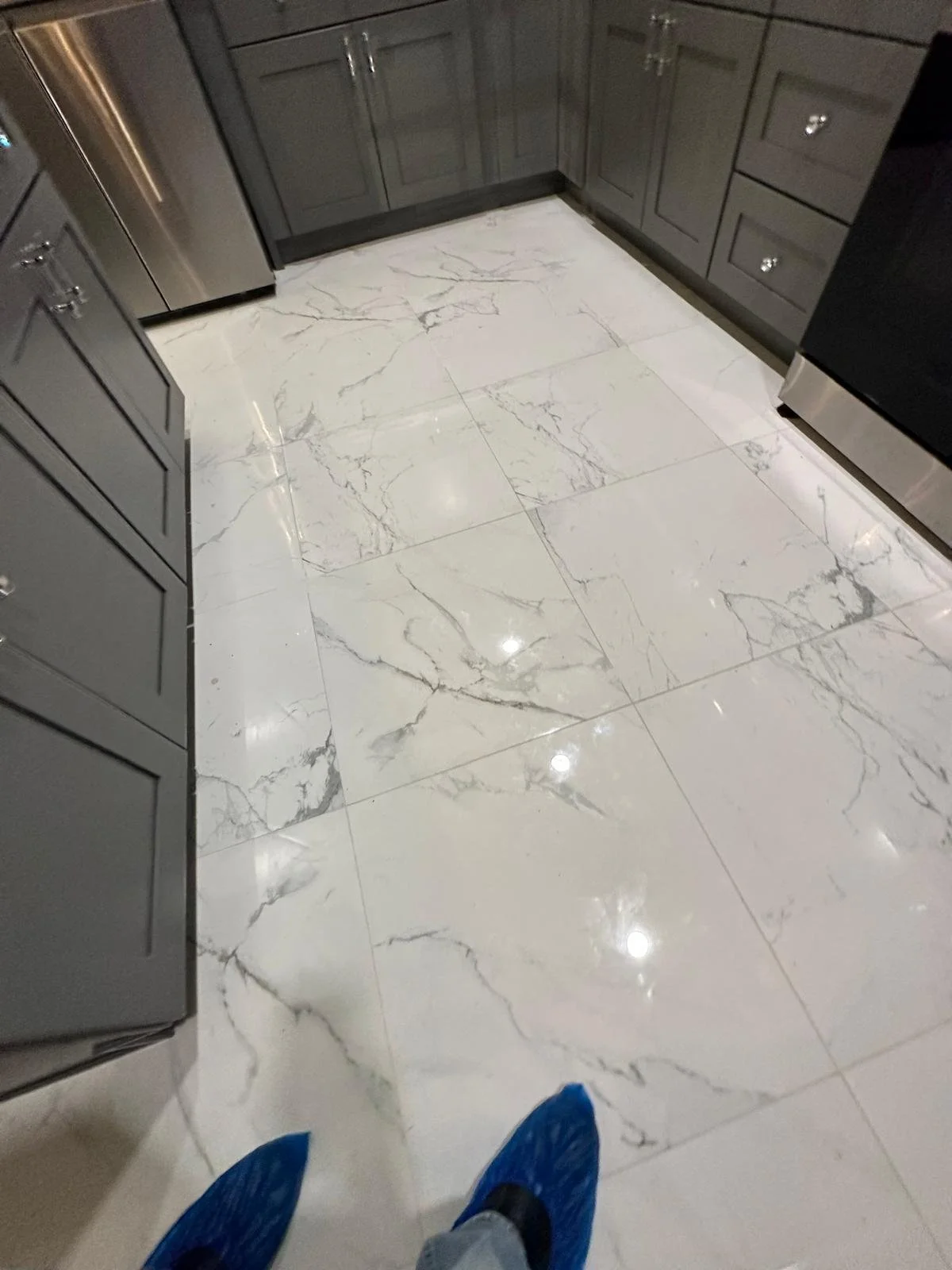 View of a modern kitchen floor with shiny white marble tiles with gray veining, gray cabinetry, and a person wearing blue slippers and gray pants.