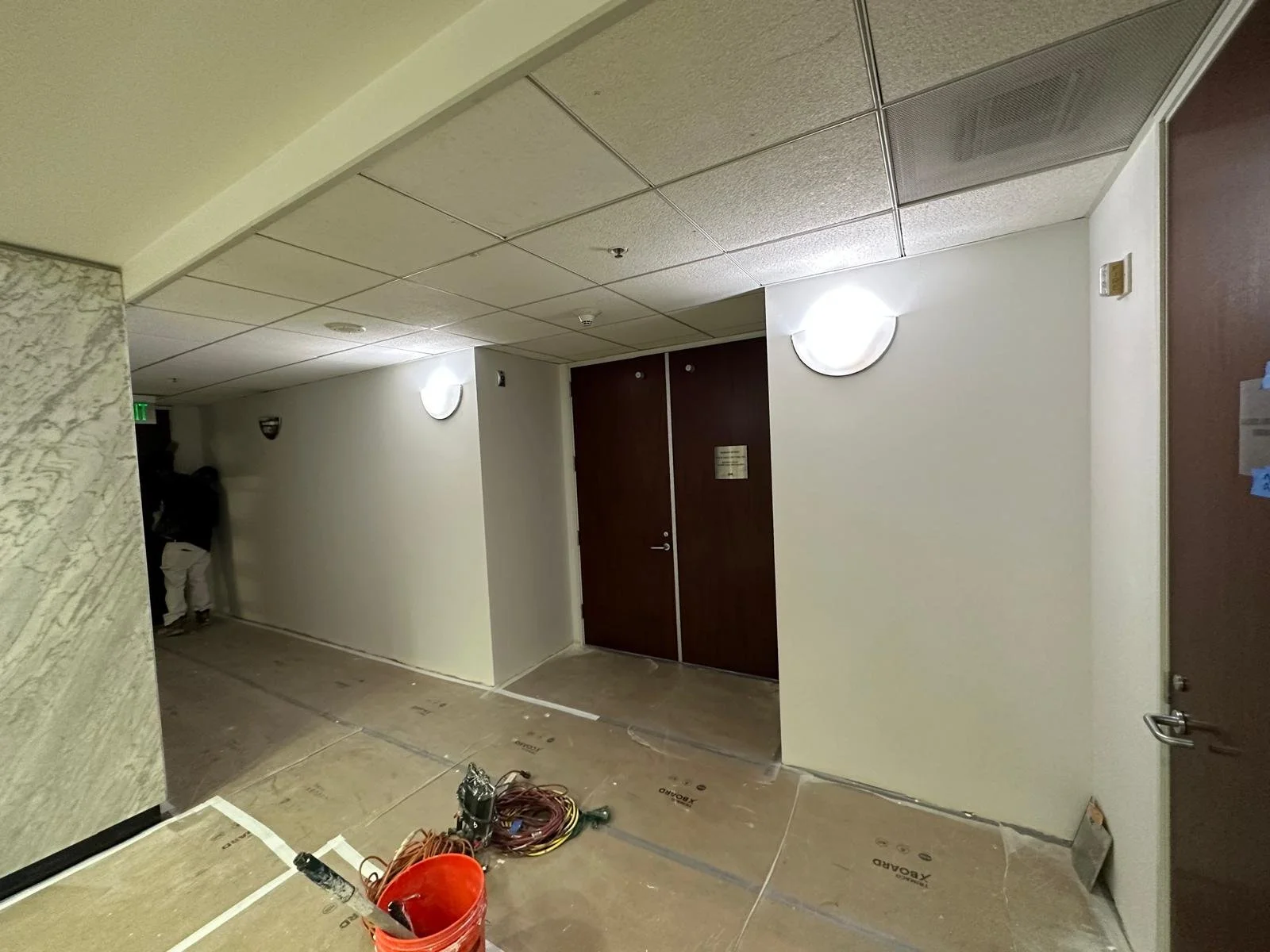An indoor corridor under construction with a partially finished ceiling, wall-mounted lights, a closed double door, and construction tools and materials on the floor.