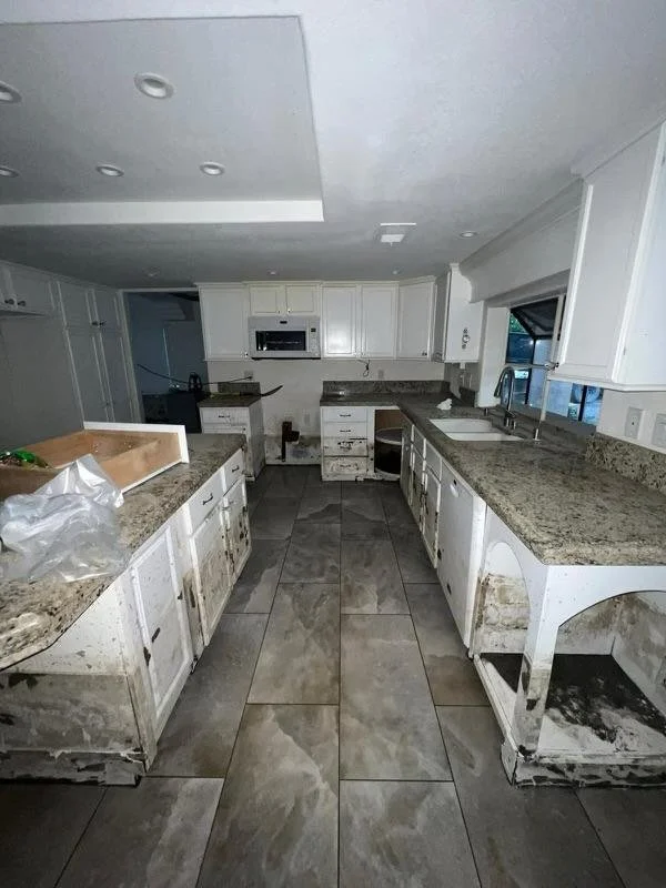 A kitchen with white cabinets and granite countertops, showing signs of damage and dirt, especially along the lower cabinets and floor.