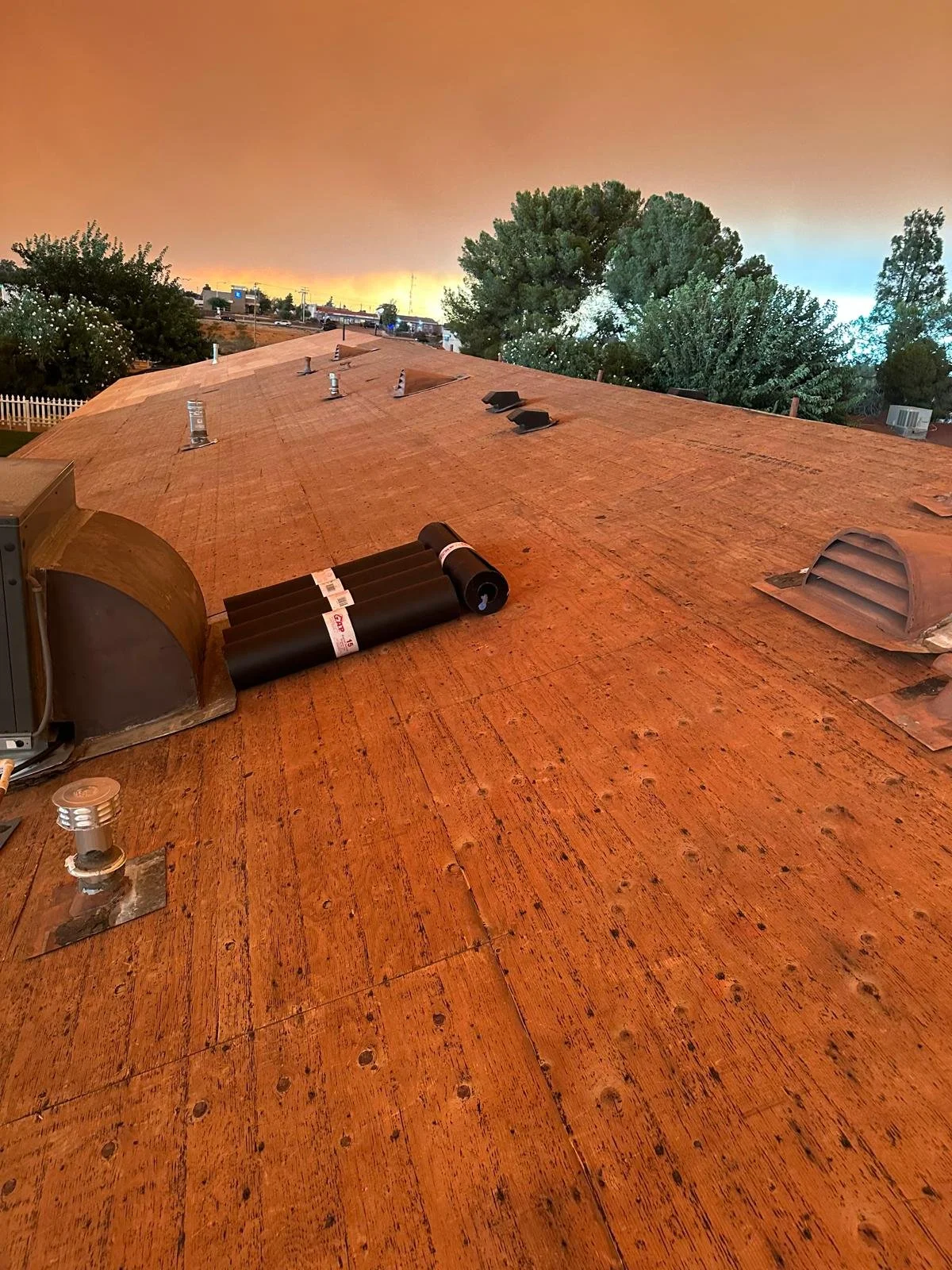 A rooftop with HVAC vents and roof materials, overlooking trees and a sunset sky with an orange hue.