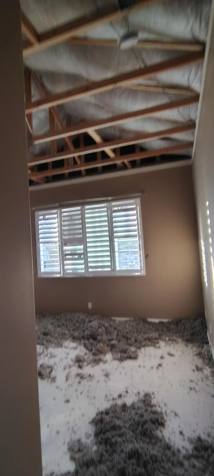 Unfinished room with exposed ceiling framing and insulation, a window with blinds, and a floor covered in dirt or debris.