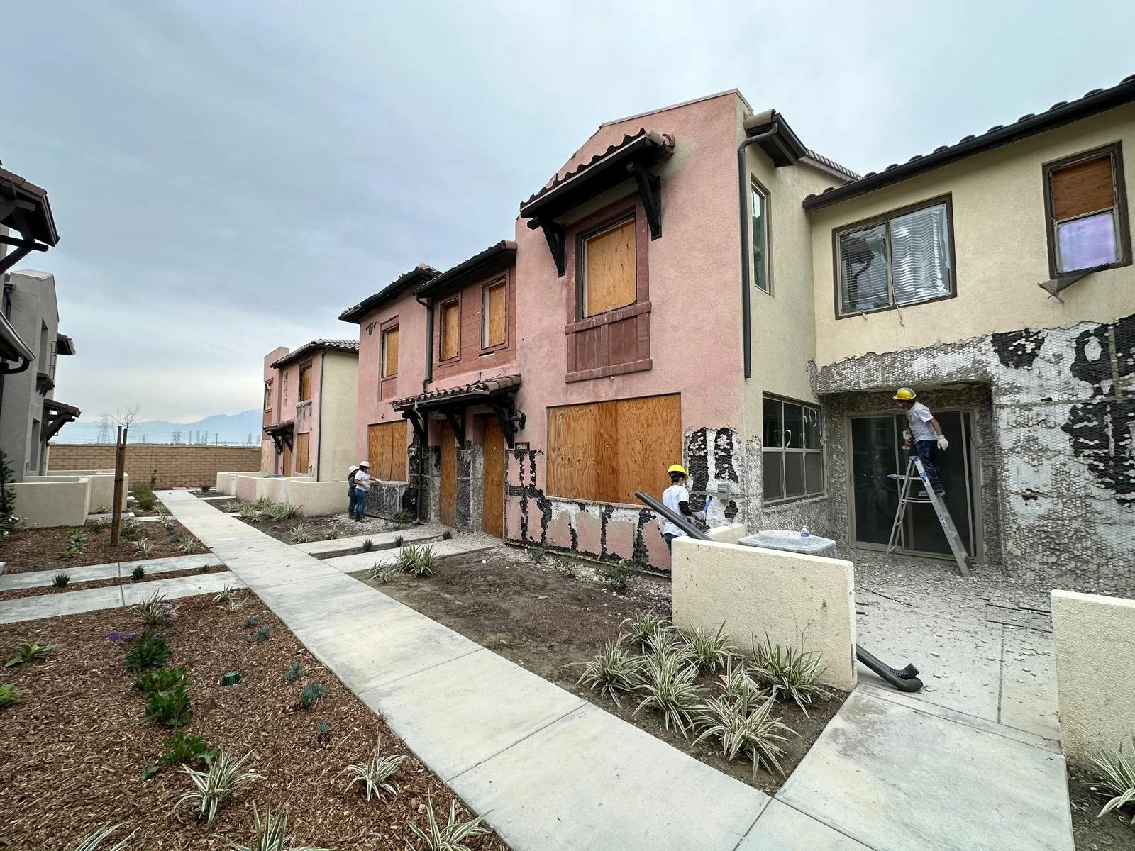 Workers with helmets working on the exterior of a multi-story residential building under renovation, with boarded-up windows and partially damaged walls, in a suburban neighborhood.