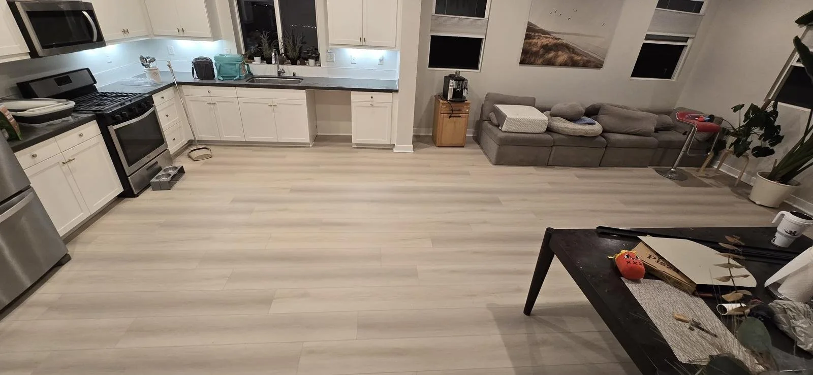 An open kitchen and living room with white cabinets, stainless steel appliances, light-colored wood flooring, a gray sofa, a coffee table with various items including a toy, and several windows and wall art.