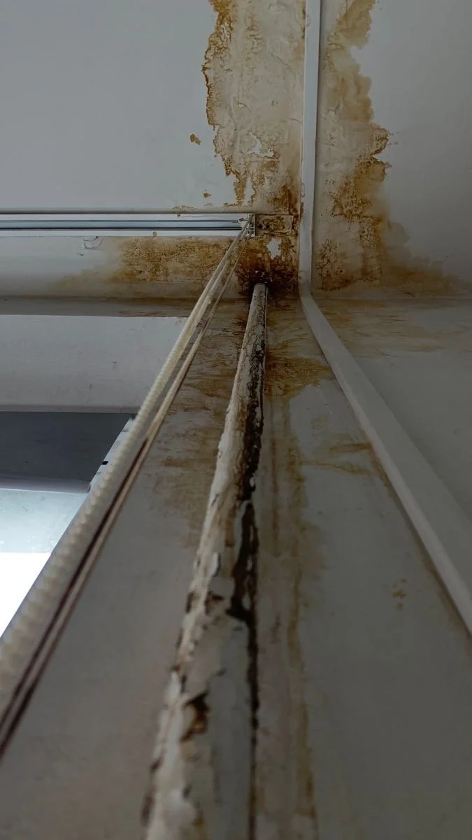 Inside a building, showing a stained and corroded corner of a ceiling and wall with rust-colored water damage and peeling paint.