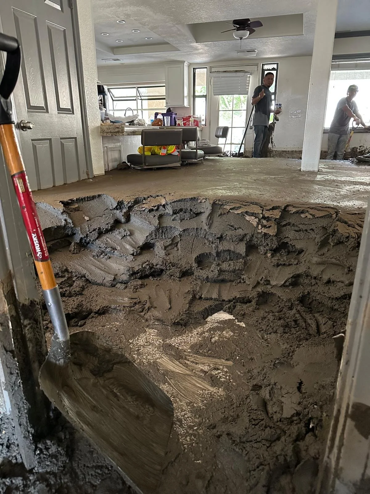 Interior of a home undergoing floor repair shows a large hole with exposed dirt and concrete, construction workers in the background, and various tools and furniture around.