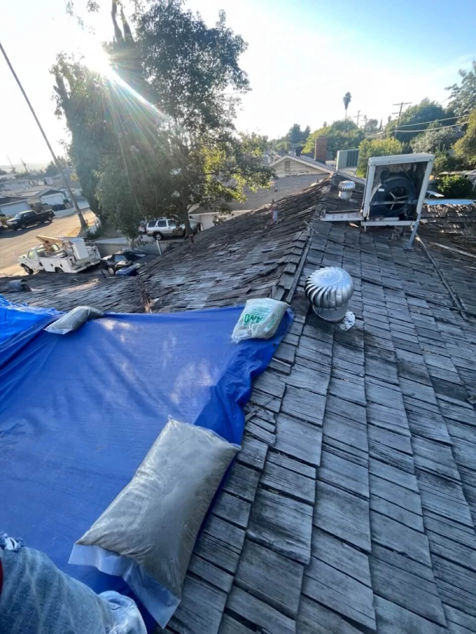 View from a rooftop showing roofing work in progress with a blue tarp, pillows, ventilation fans, and nearby streets and houses.