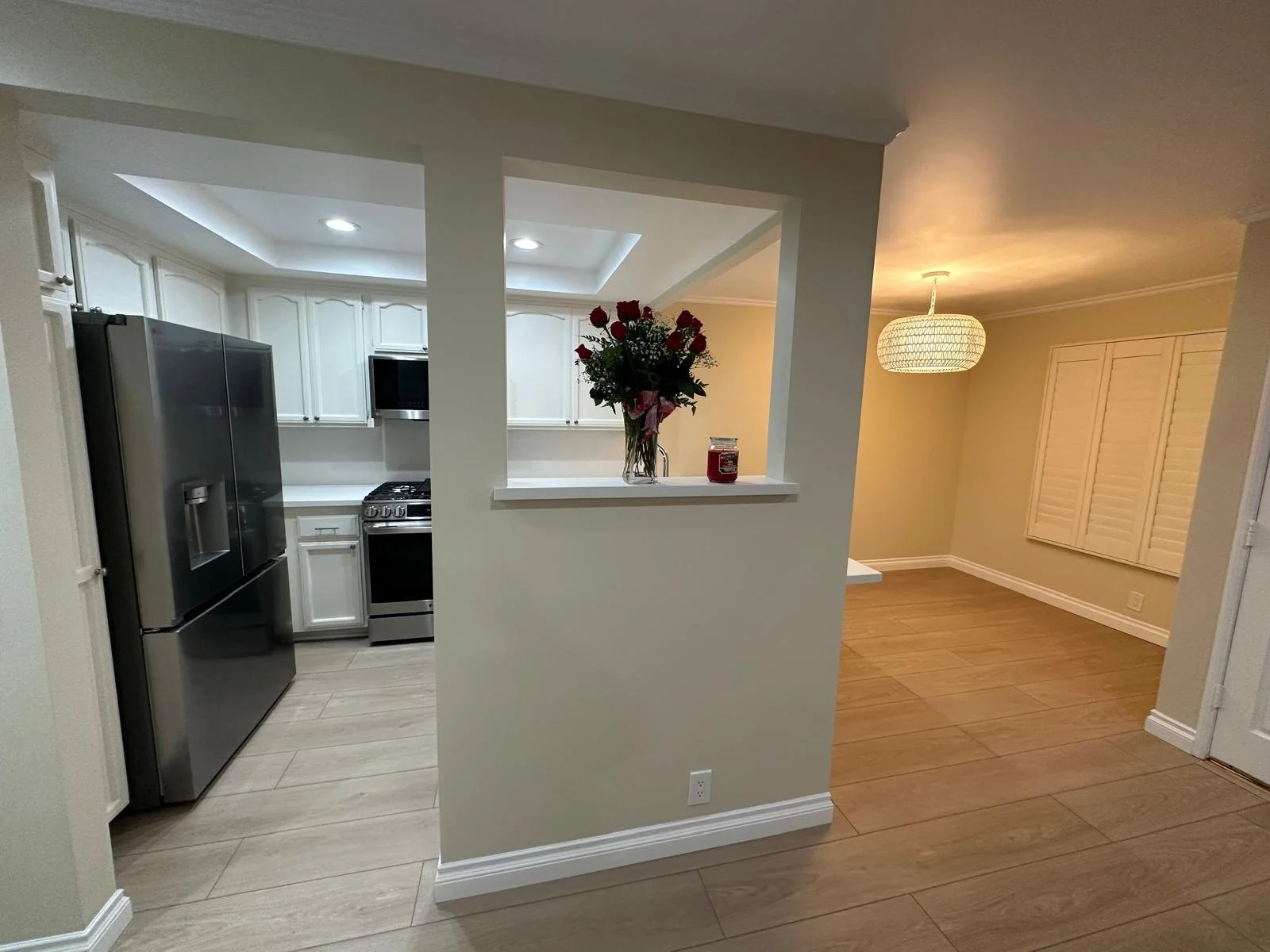 Interior view of a kitchen with white cabinets, black appliances, and a pass-through window with a flower vase on the ledge. Adjacent room with a ceiling light and a closed shutter window