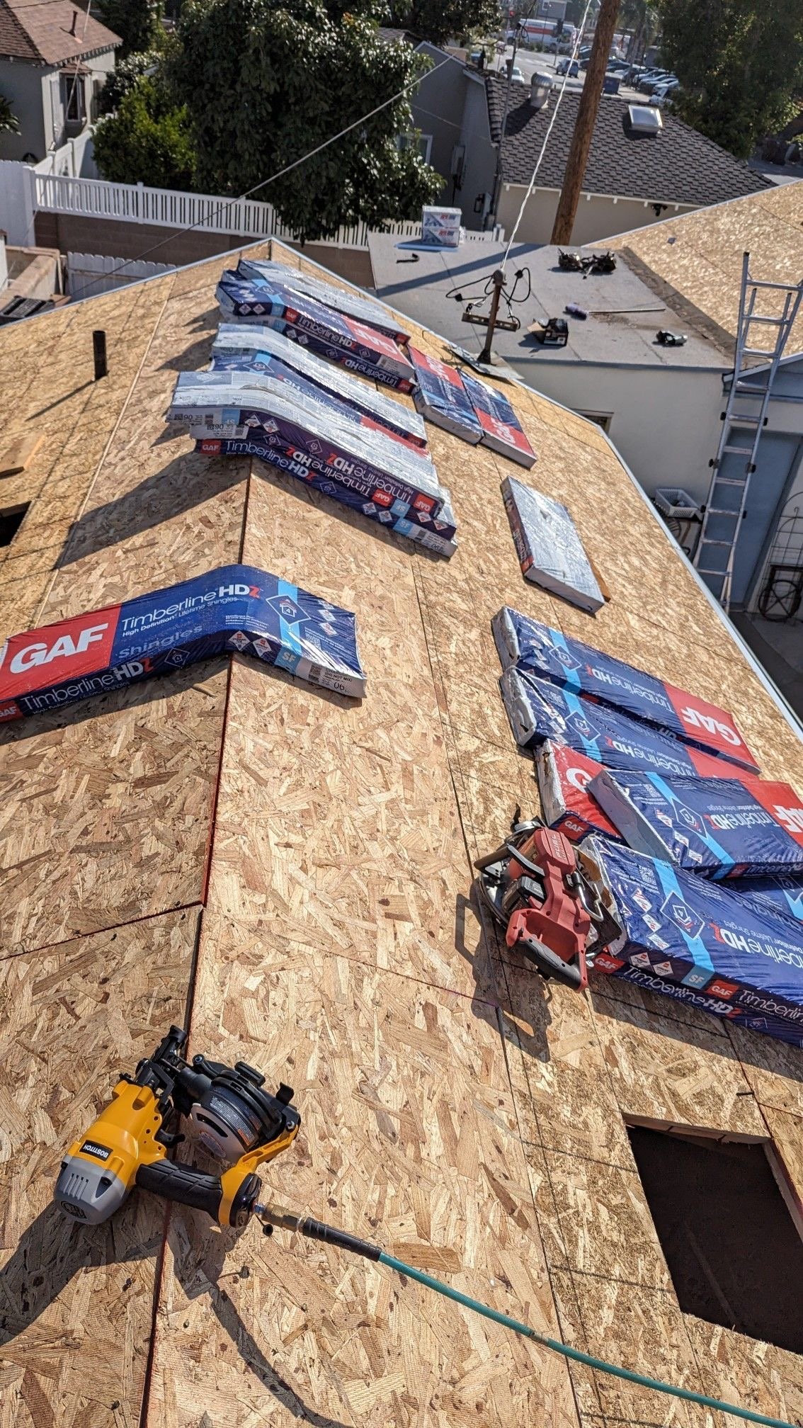 View of a rooftop under construction with sheets of oriented strand board (OSB) sheathing, a nail gun, and bundled roofing materials labeled Timberline HD, during daytime. In the background, neighboring rooftops, trees, power lines, and a distant par