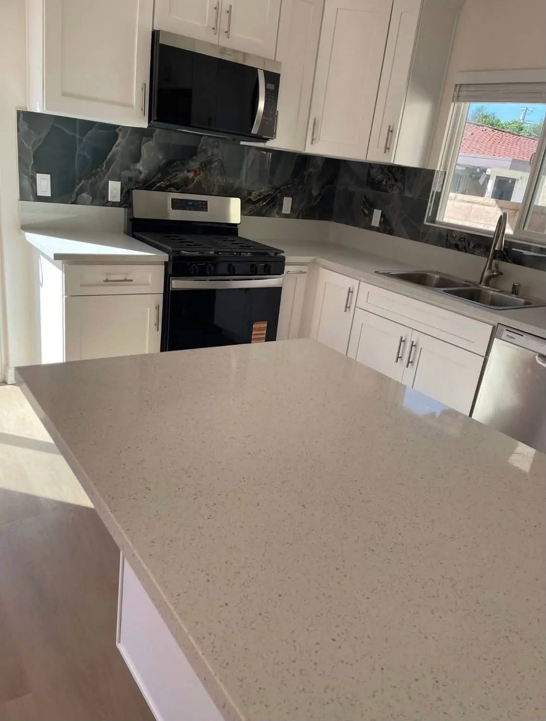 Kitchen with white cabinets, black and gray marble backsplash, stove, microwave, double sink, window, and kitchen island.
