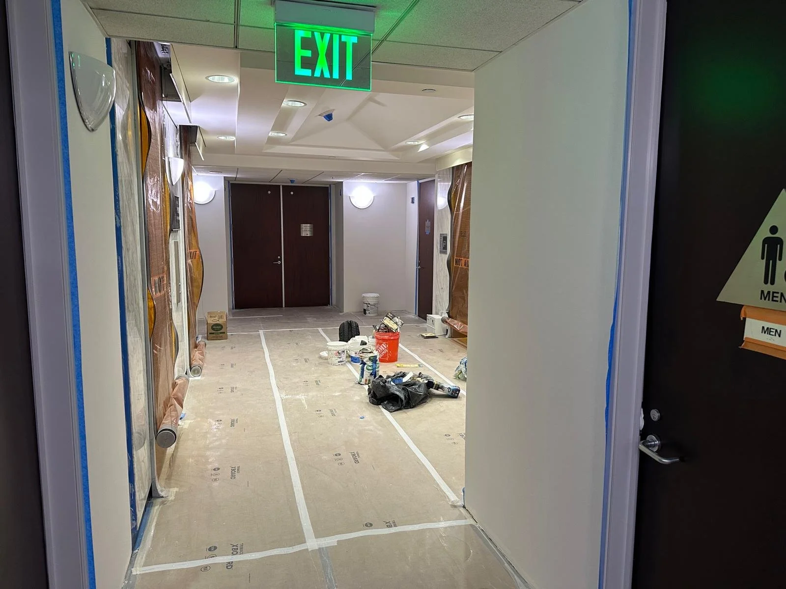 An underground corridor under construction with various construction materials, tools, and equipment on the floor. There are two closed doors at the end of the corridor and a green emergency exit sign hanging from the ceiling.