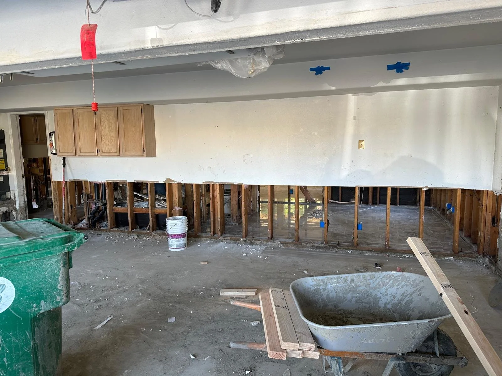 Interior view of a kitchen under renovation with exposed wall studs, wooden cabinets, construction tools, a wheelbarrow, and debris on the floor.