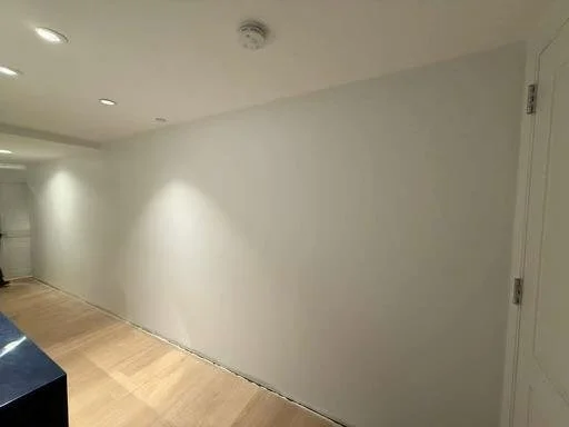 Empty white wall in a room with a closed white door on the right and ceiling lights