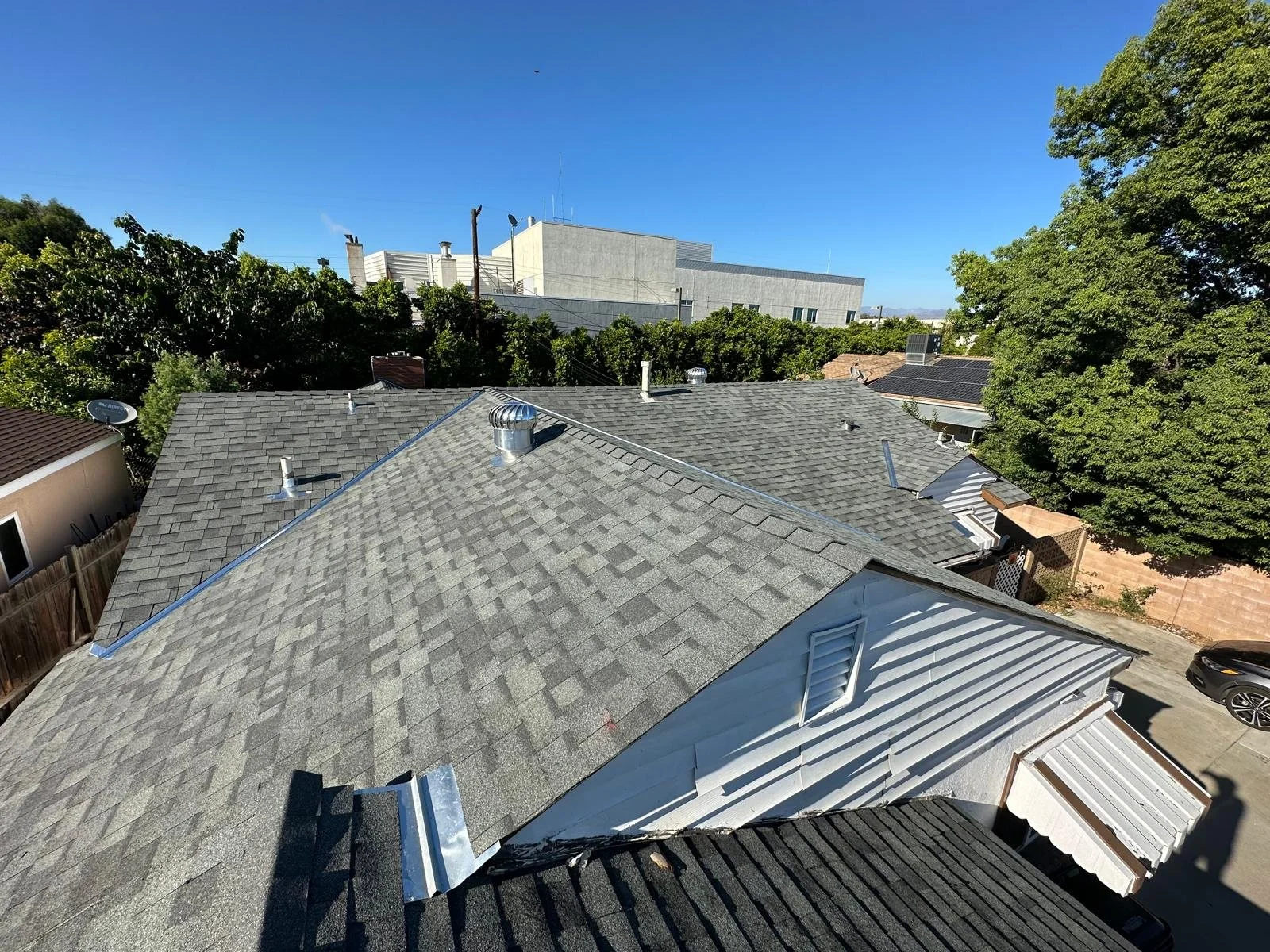 A view of rooftops from above, showing gray shingle roofs, vents, and a white house with a metal roof, with trees and a clear blue sky in the background.