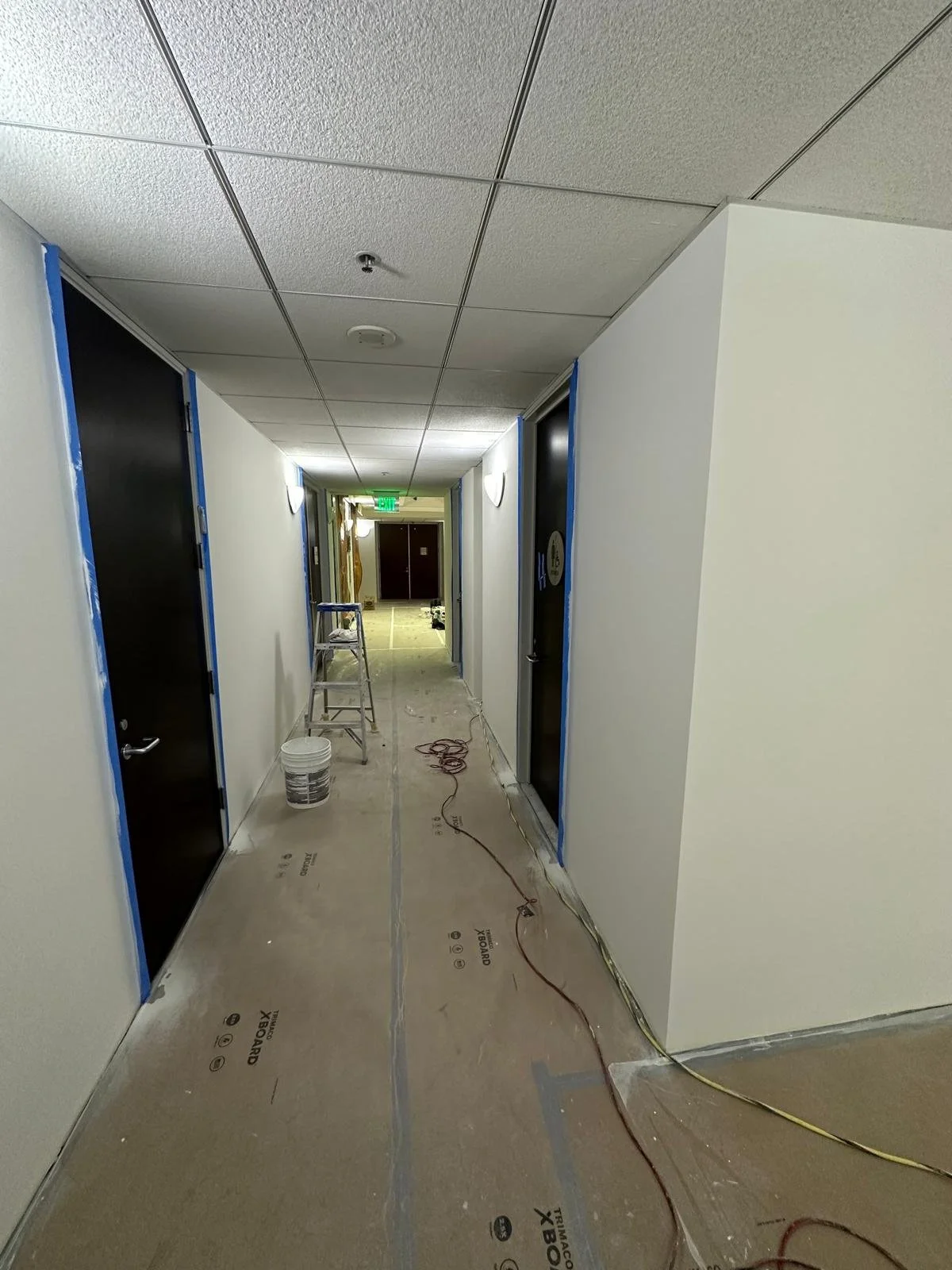 A hallway under construction with black doorways on both sides, a ladder, and construction materials on the floor.