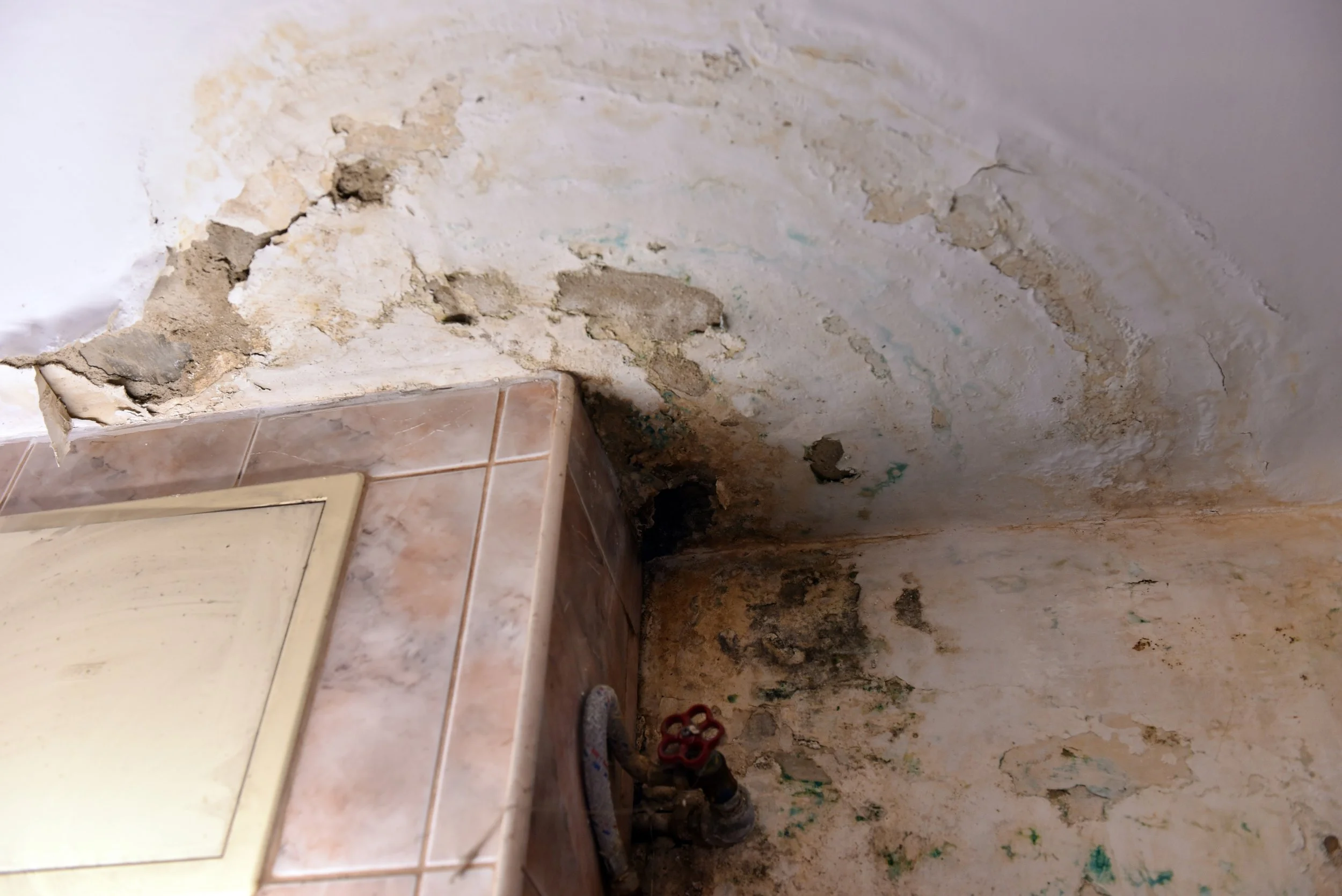A section of damaged or deteriorating wall with visible cracks and peeling paint near a tiled step and plumbing pipes.