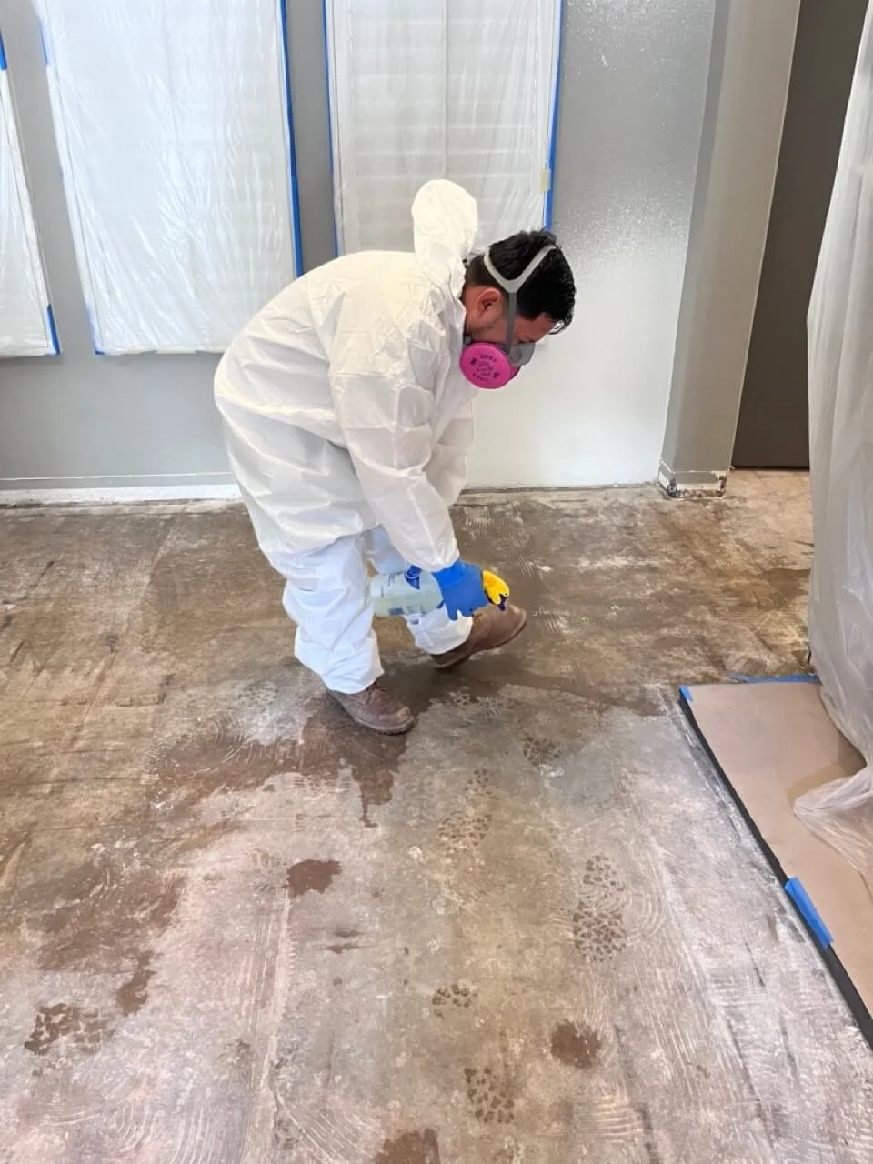 A person wearing protective gear, including a white suit, a pink respirator mask, gloves, and boots, is disinfecting or cleaning a concrete floor in a construction or renovation site.