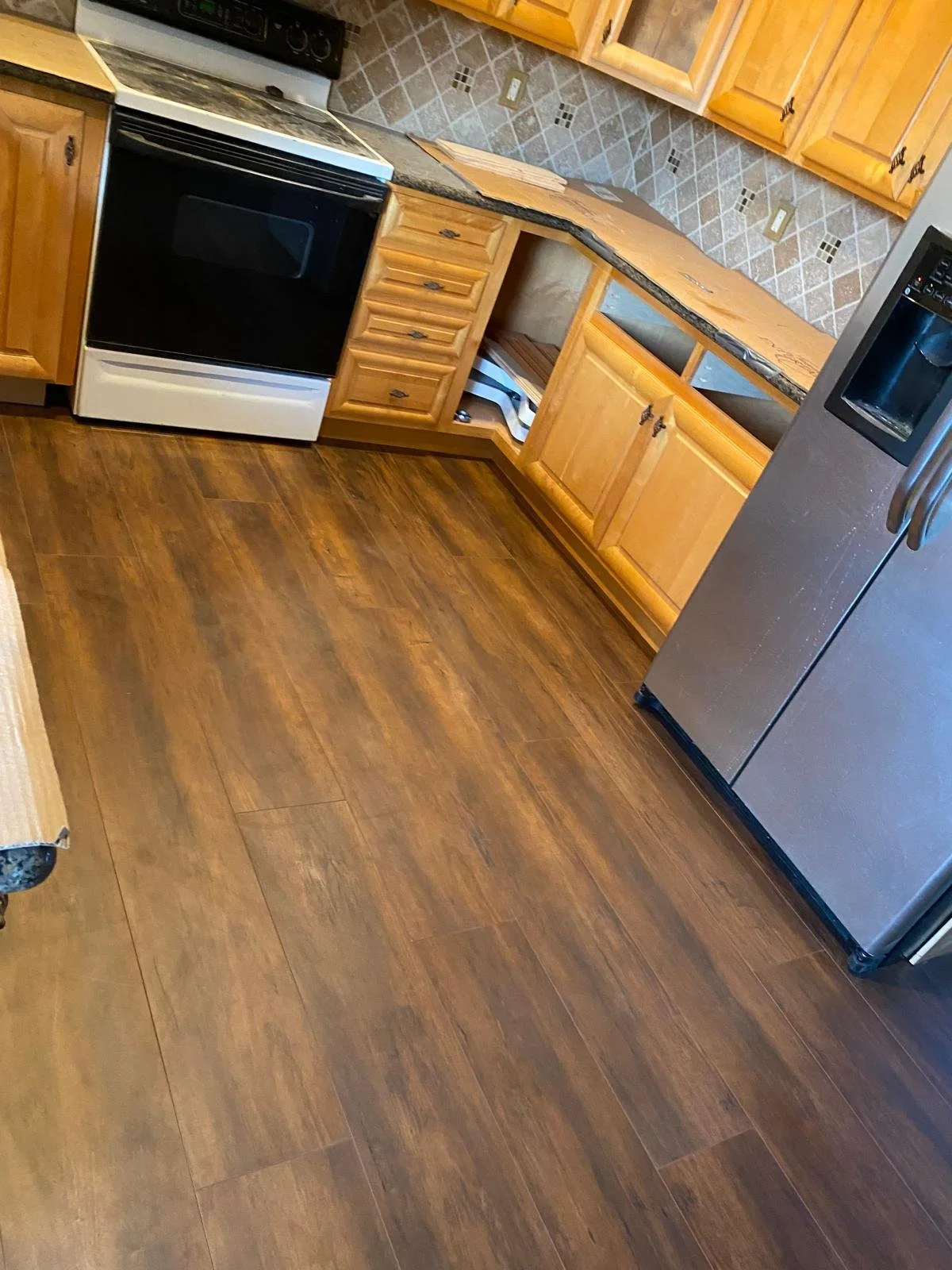 A kitchen with wooden cabinets, a stainless steel refrigerator, and a black and white stove. The floor is hardwood, and the countertop has a granite surface with a tiled backsplash.