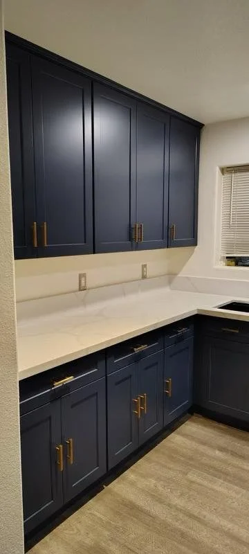 Photo of a kitchen with navy blue cabinets and white countertops, located beneath a window and above a light-colored wood floor.