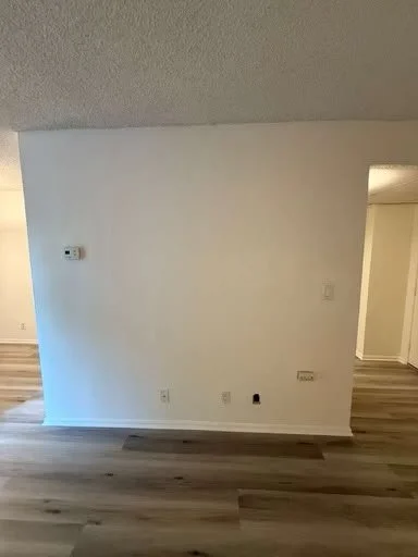 Empty white wall in a room with a thermostat, electrical outlets, and a power outlet, with laminate wood flooring and an adjacent hallway.