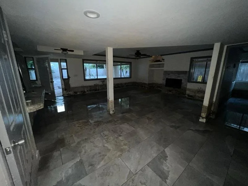 Empty living room with wet floor, damaged walls, and multiple windows showing an outdoor yard.