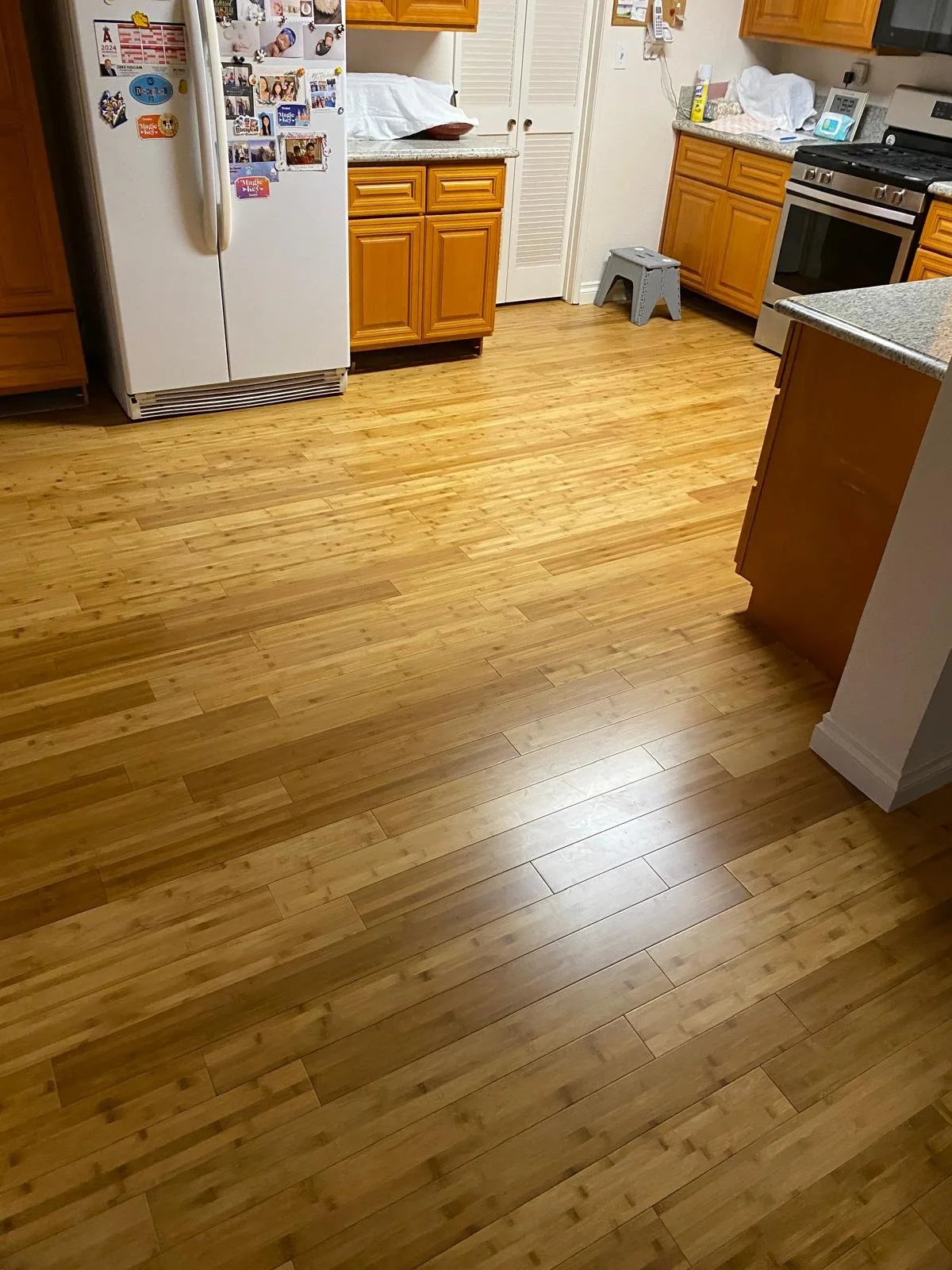 Empty wooden kitchen floor with visible floorboards, kitchen cabinets, a white refrigerator with magnets, kitchen countertops, stove, small step stool, and some kitchen appliances and decor.